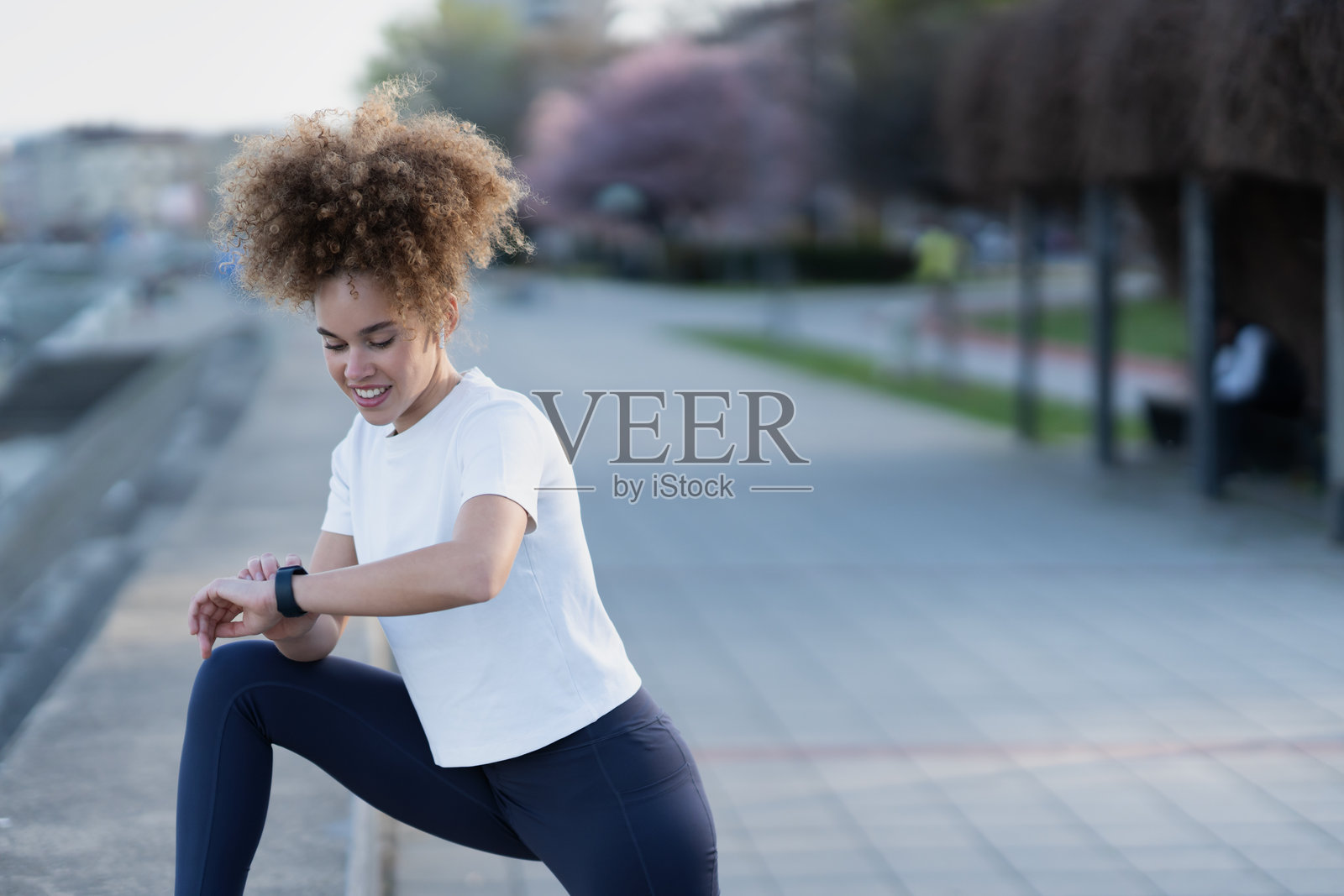 一位年轻女性穿着运动服，在水边伸展身体时查看她的智能手表。她微笑着，享受着在城市环境中与树木和城市景观相伴的健身时光。照片摄影图片