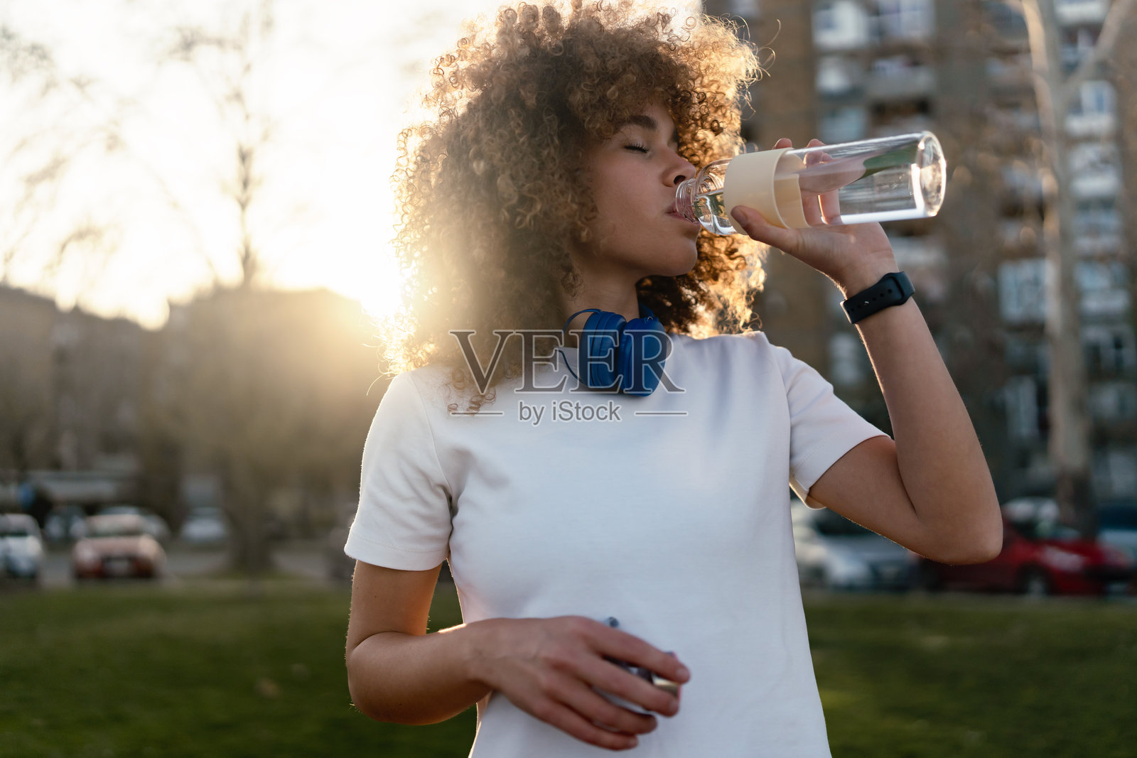 一位年轻的卷发女性在城市的日落时分户外站着，喝着玻璃瓶中的水。她脖子上挂着无线耳机，手腕上戴着智能手表，展现出健康的生活方式。照片摄影图片