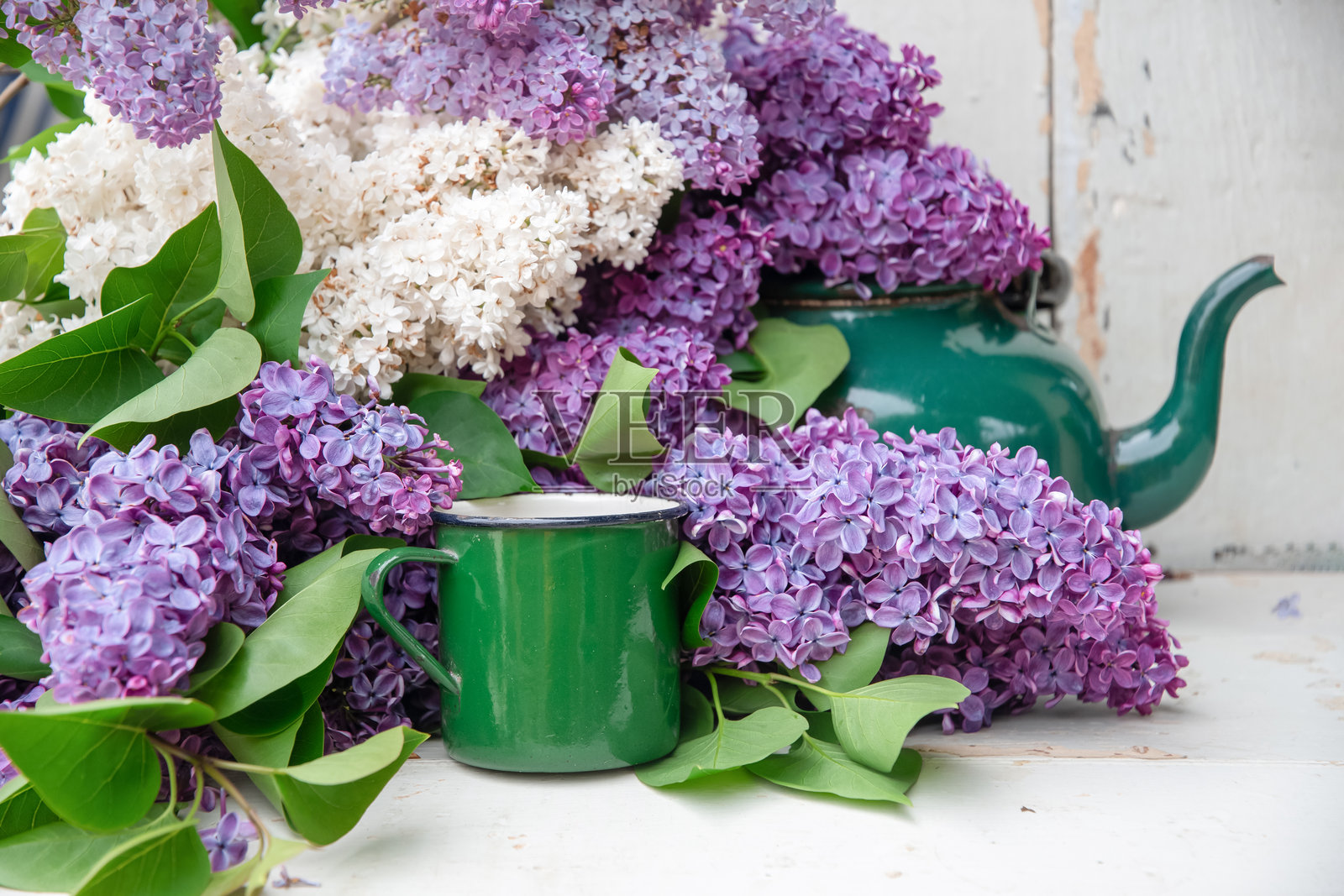 一大束不同色调的紫丁香围绕着一个绿色搪瓷杯和茶壶，摆放在白色的乡村风格桌子上，营造出迷人的静物场景。照片摄影图片