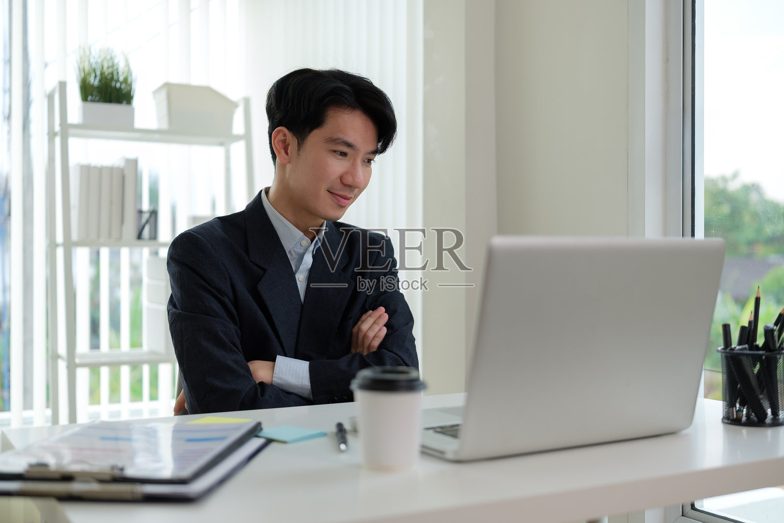 身穿正式服装的商人坐在白色桌子前，双臂交叉，满意地看着他的笔记本电脑。照片摄影图片