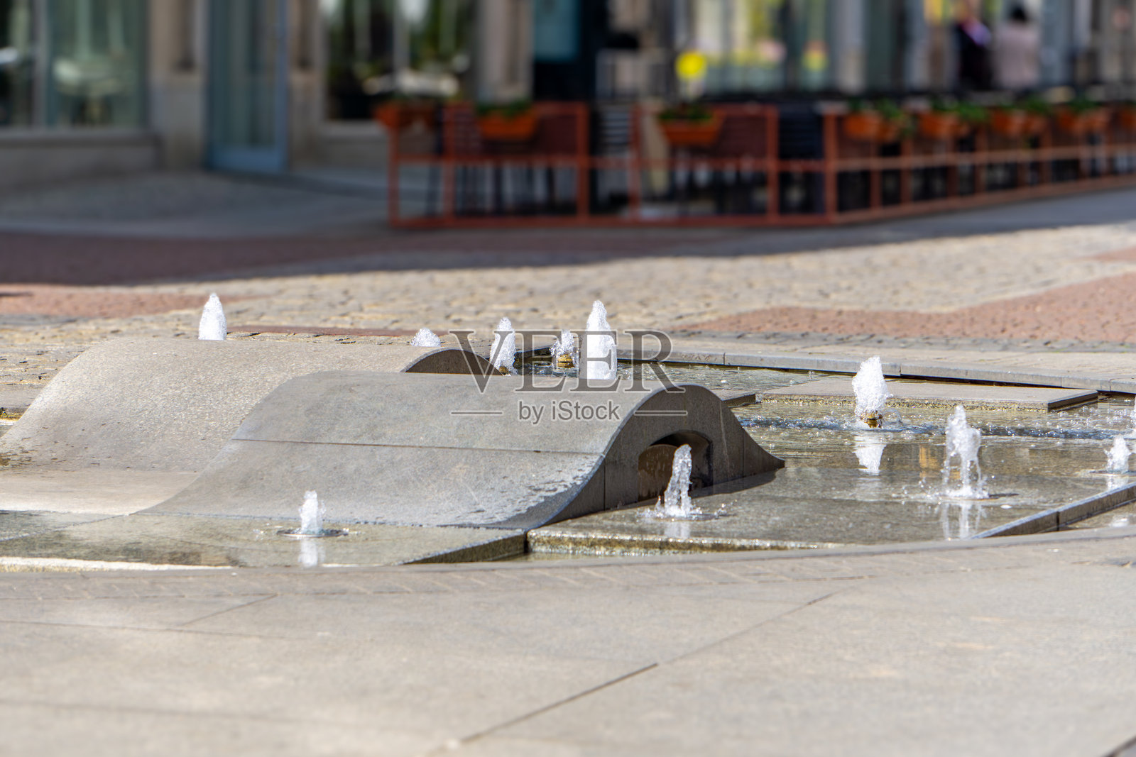 一个现代设计的城市喷泉，水从混凝土装置的多个点流出，位于市中心的步行街上。背景中可见咖啡桌和商店橱窗。照片摄影图片