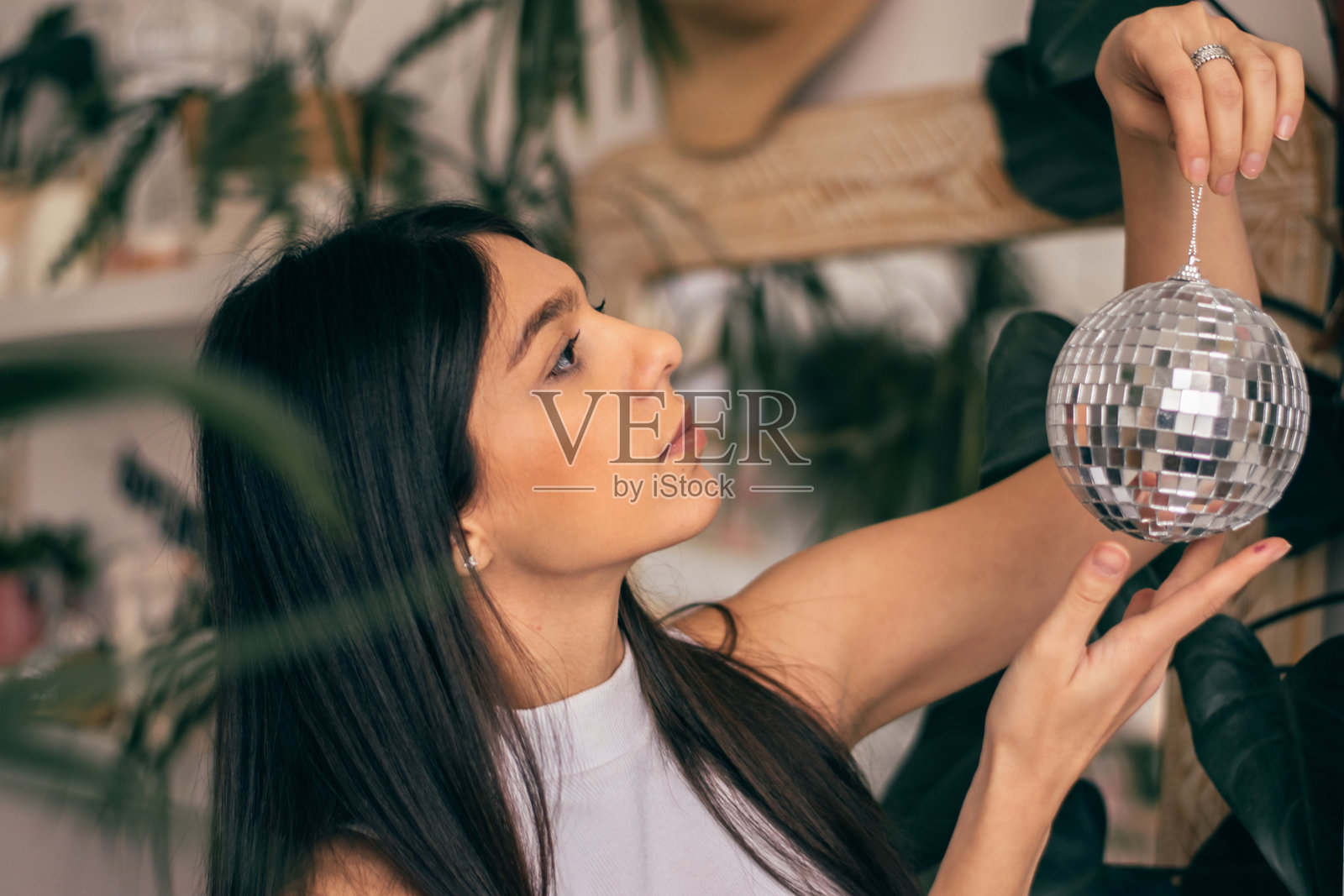 一位年轻的深发女性手中拿着一个镜面迪斯科球。她在一个有室内植物的房间里。生物亲和力概念。照片摄影图片