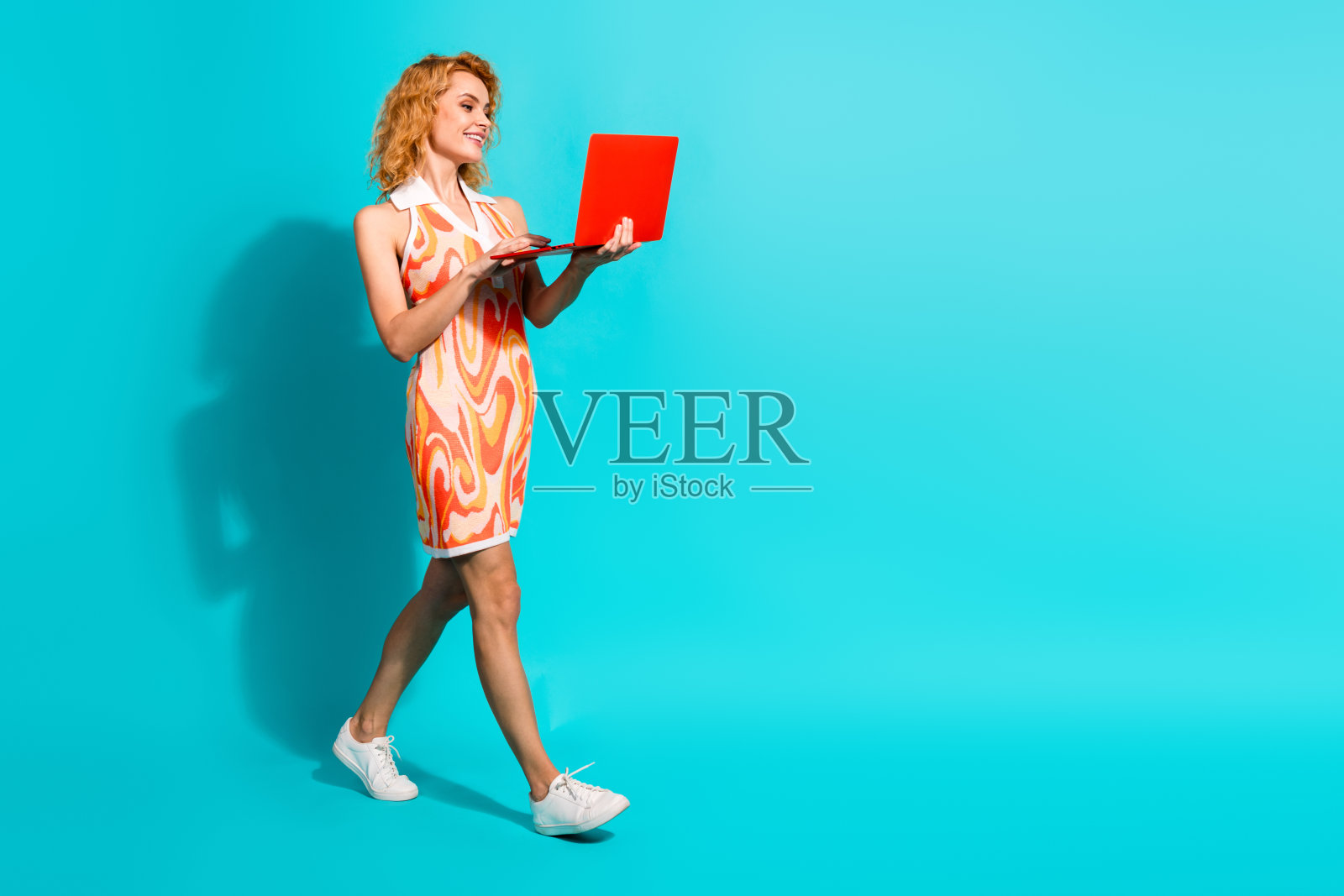 时尚年轻女性穿着色彩斑斓的裙子，坐在鲜艳的青色背景前，使用红色笔记本电脑阅读，散发出自信与魅力。照片摄影图片