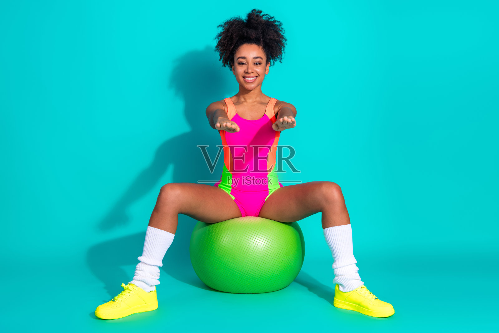 穿着鲜艳运动服的快乐年轻女性在绿健身球上锻炼，背景为青绿色，展现出时尚自信的积极健康生活方式。照片摄影图片