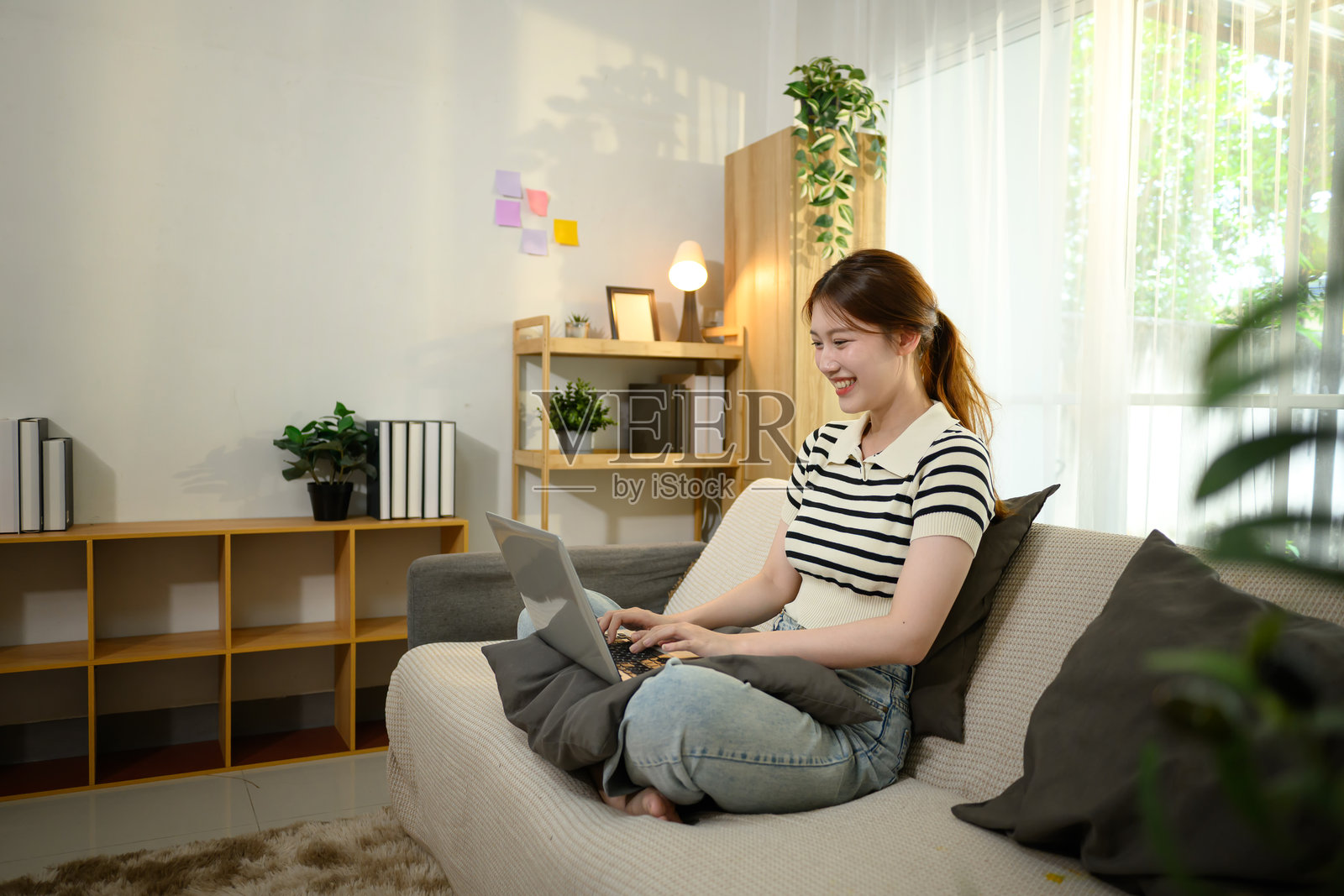 快乐的亚洲女性坐在沙发上，在明亮的房间里使用笔记本电脑照片摄影图片