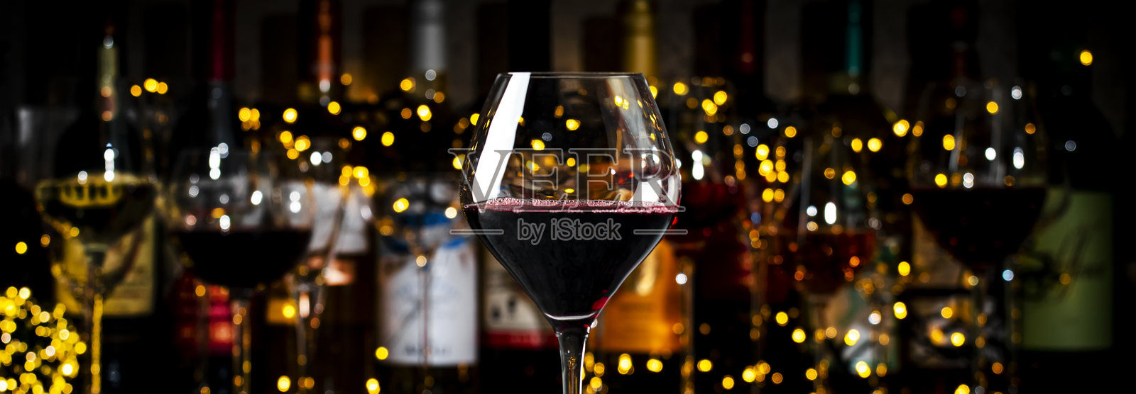 红色梅洛葡萄酒杯和酒瓶在昏暗酒吧背景下，灯光闪烁。照片摄影图片