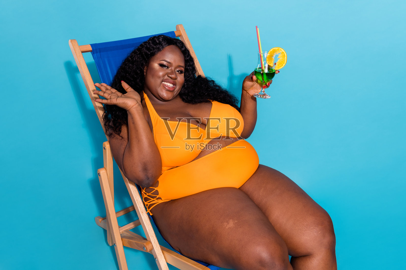 曲线优美的女士坐着享受炎热的天气，手持玻璃杯，向大家挥手，每个人都是美丽的，背景为蓝色。照片摄影图片
