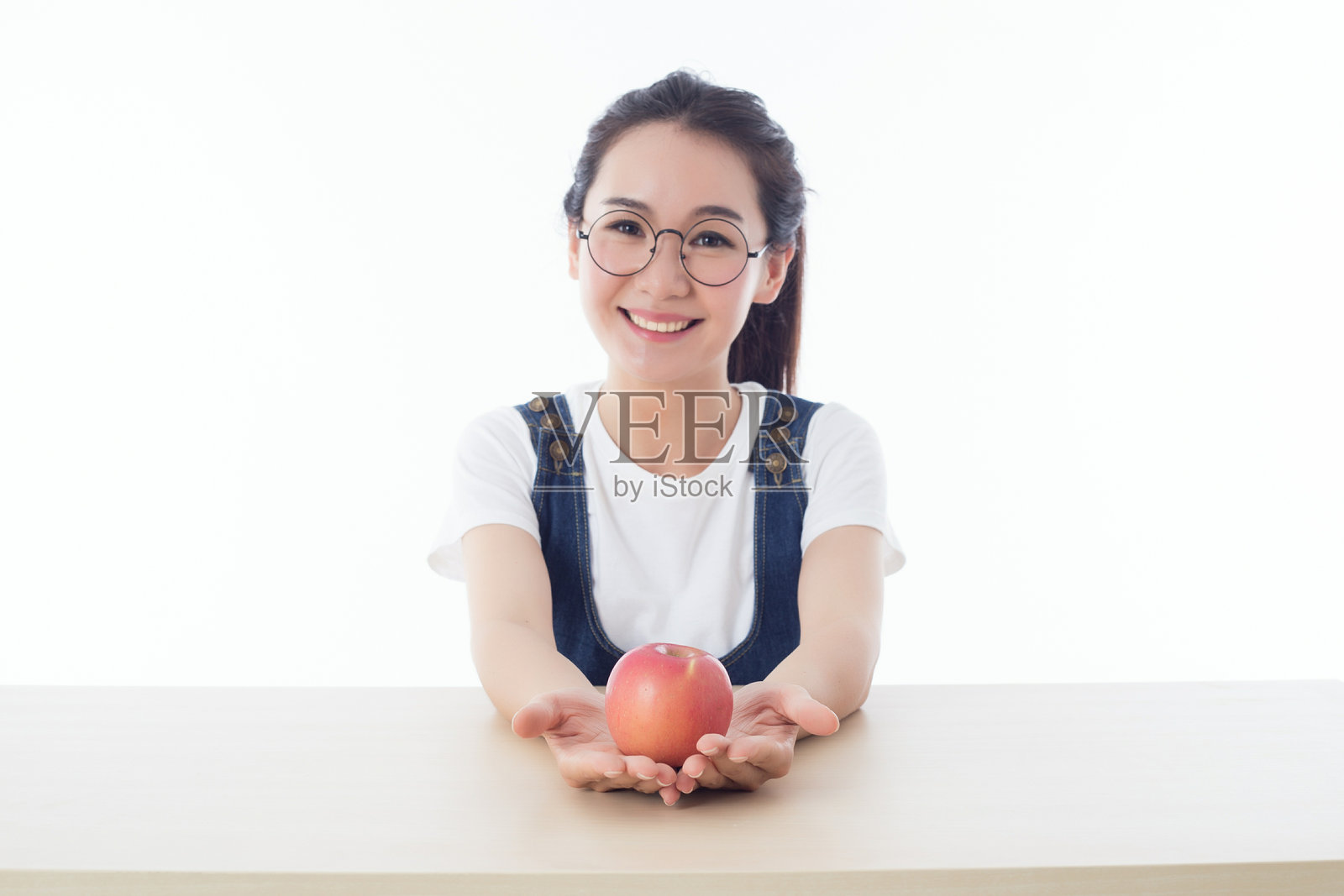 女孩坐在白色背景的桌子旁，手里拿着一个苹果。照片摄影图片