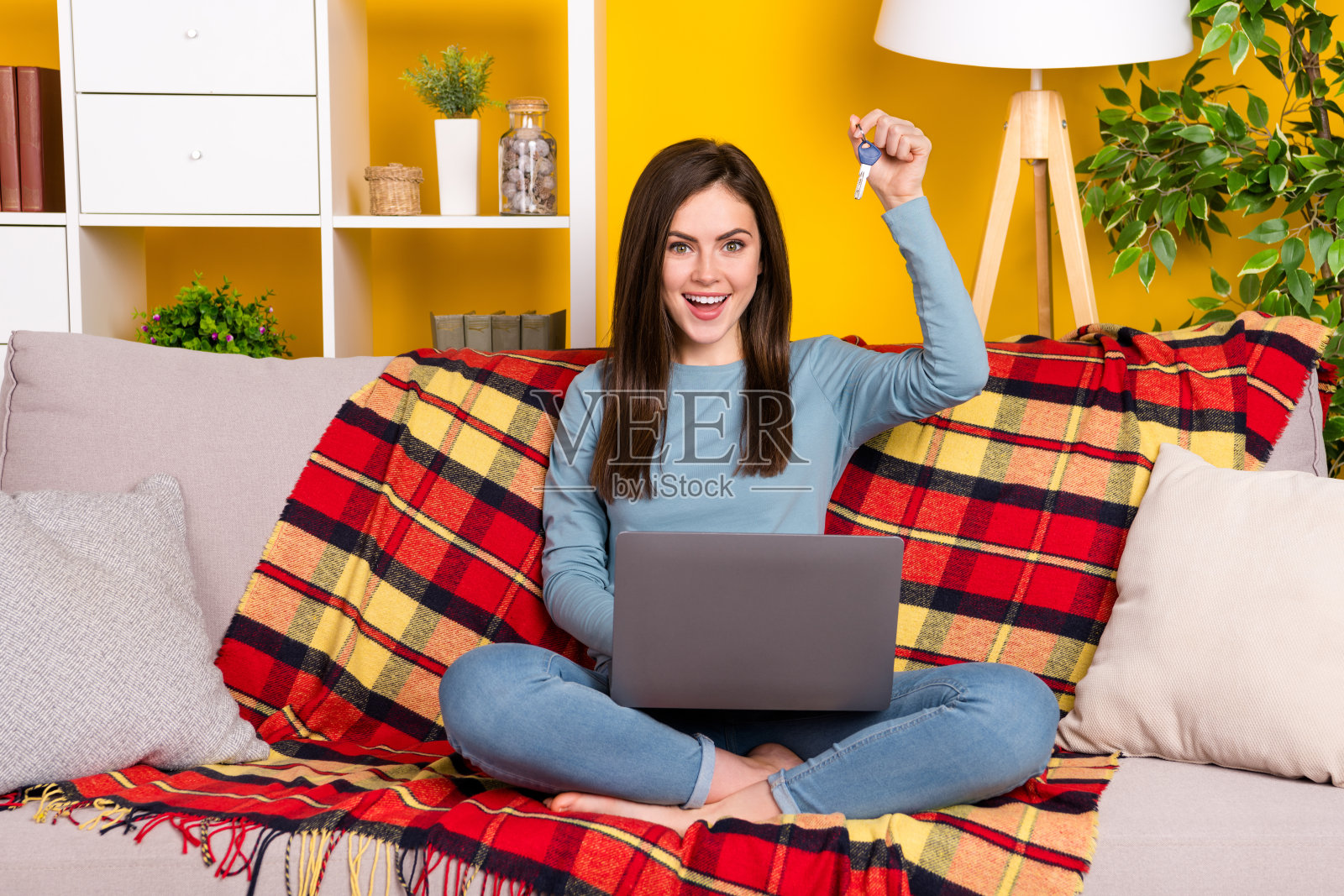 全身肖像的迷人女士赤脚坐在沙发上，手持钥匙，背景为黄色。照片摄影图片