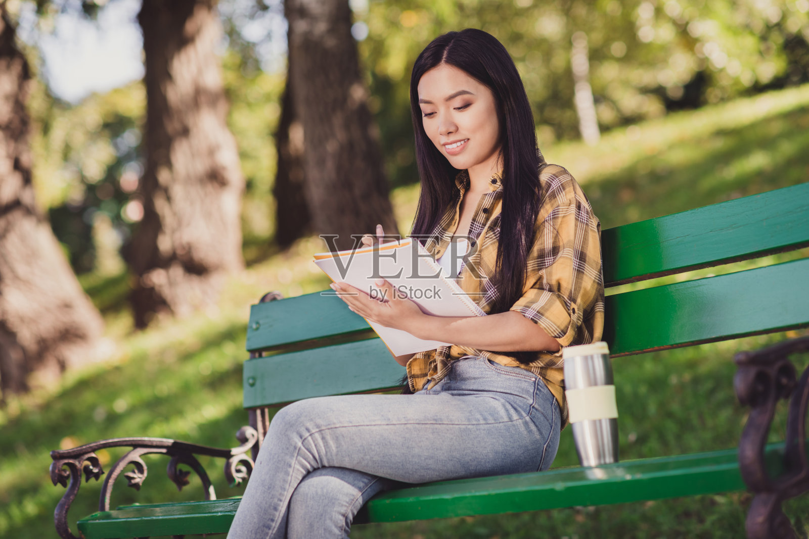 穿着格子衬衫的可爱年轻女士在城市公园的长椅上微笑着写书。照片摄影图片