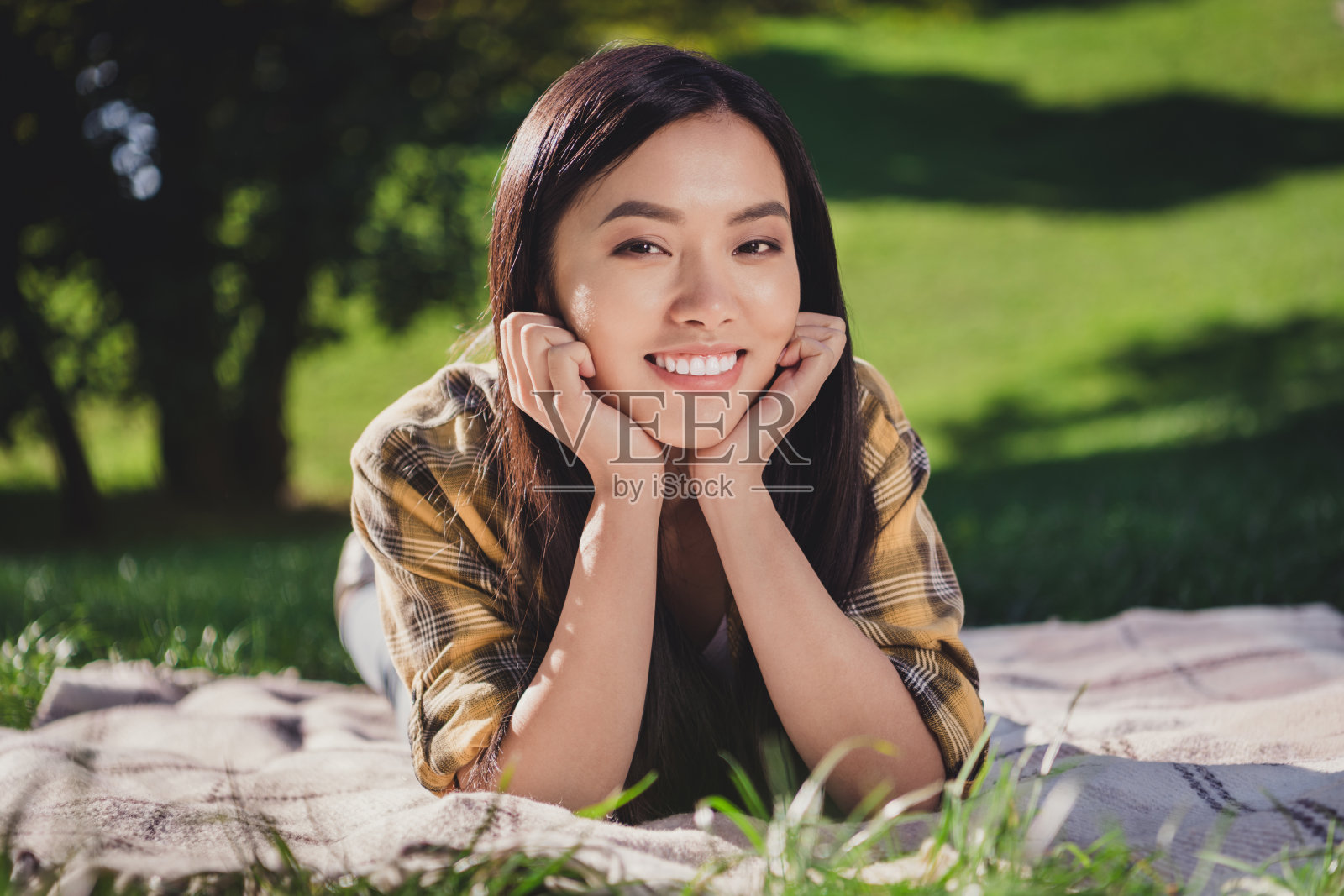 阳光下穿着格子衬衫的漂亮年轻女士微笑着躺在城市公园的绿色草地上。照片摄影图片