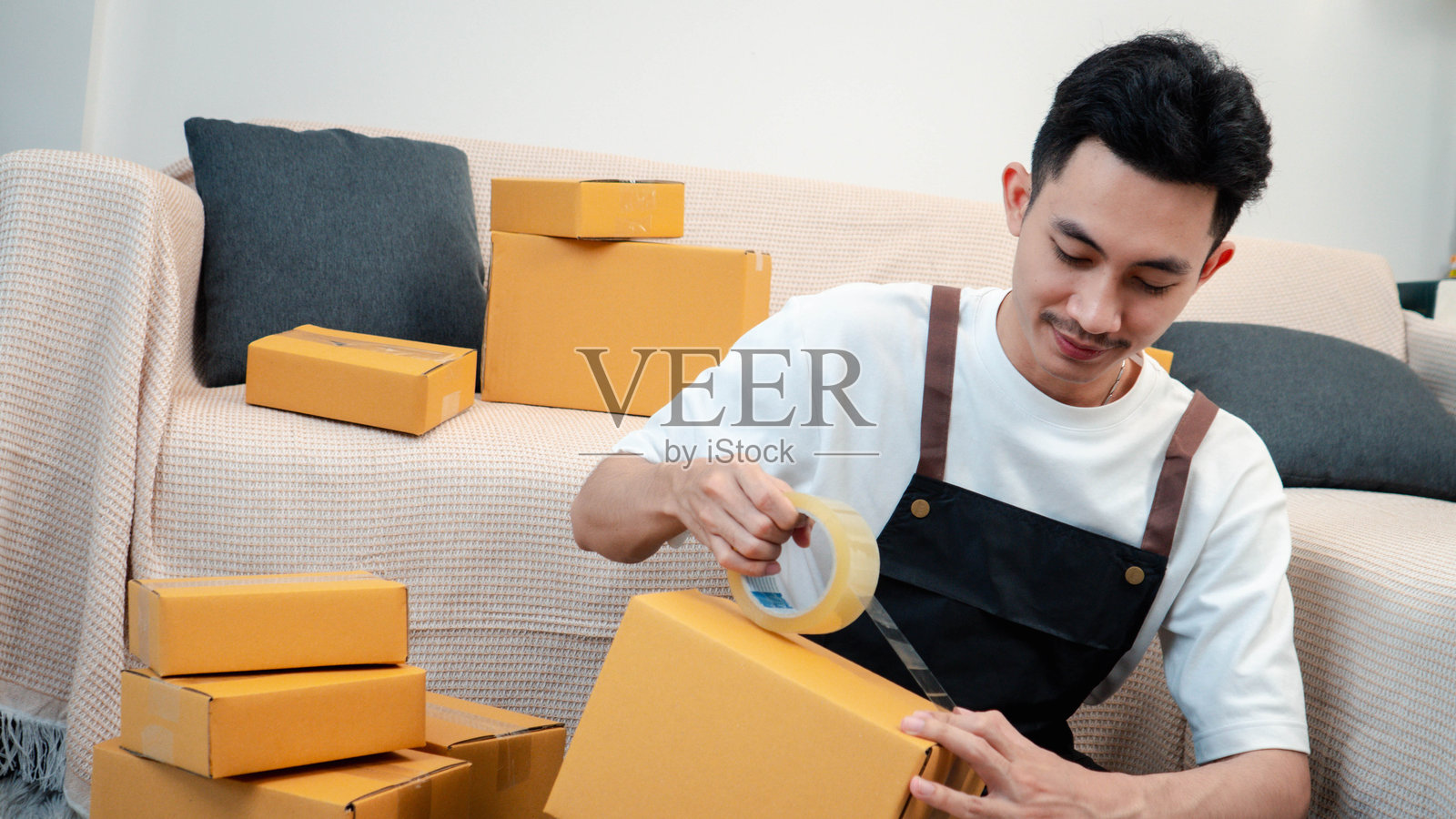 男性使用透明胶带将包裹箱固定，准备在家进行商品包装，为向客户销售的盒子打包，体现了在线购物的概念。照片摄影图片