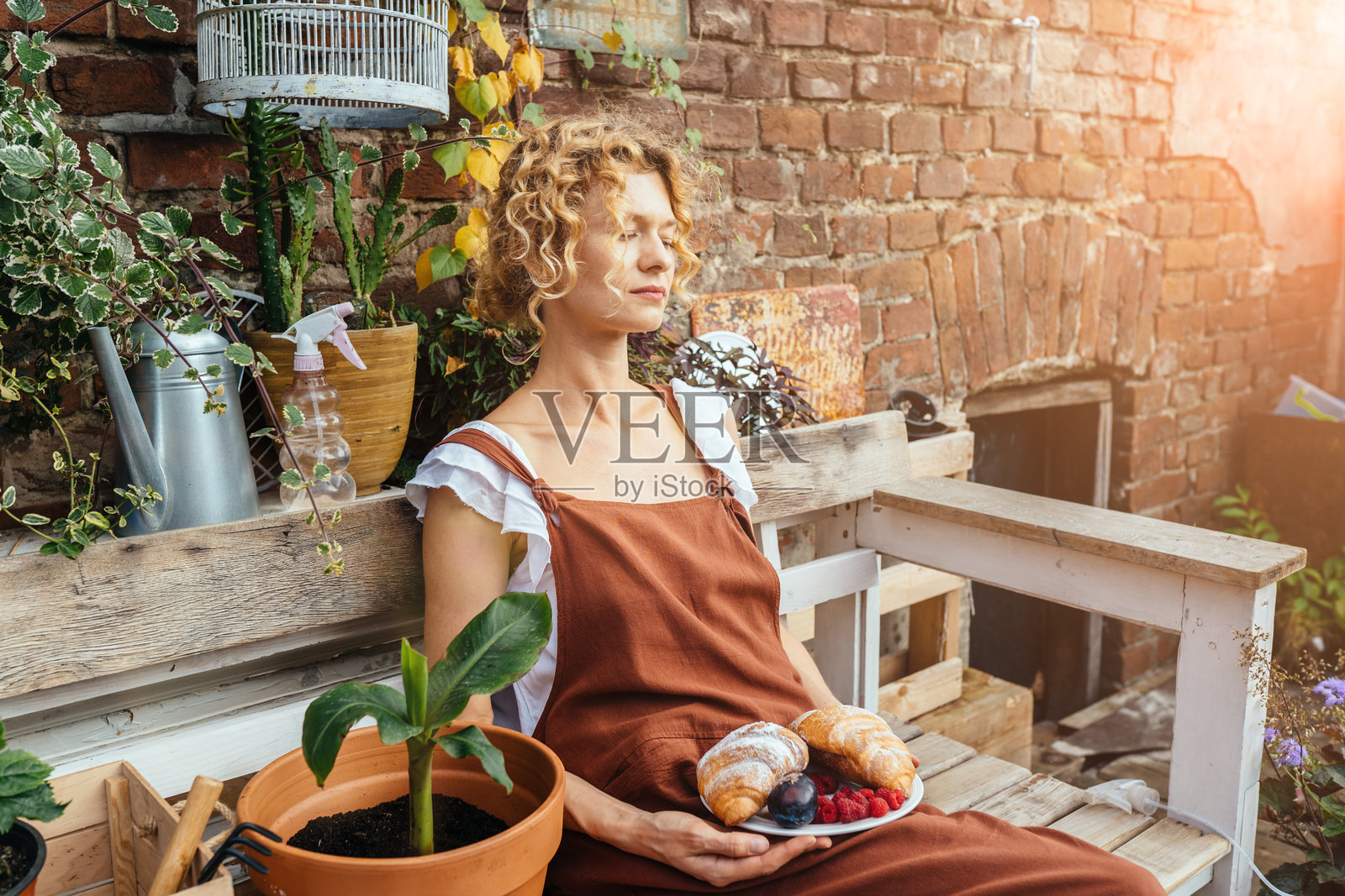 中年疲惫的园丁穿着棕色工装，在花园里工作后放松，手里拿着一盘可颂，坐在老旧的长椅上，靠在古老的砖墙上。休闲，园艺概念。照片摄影图片