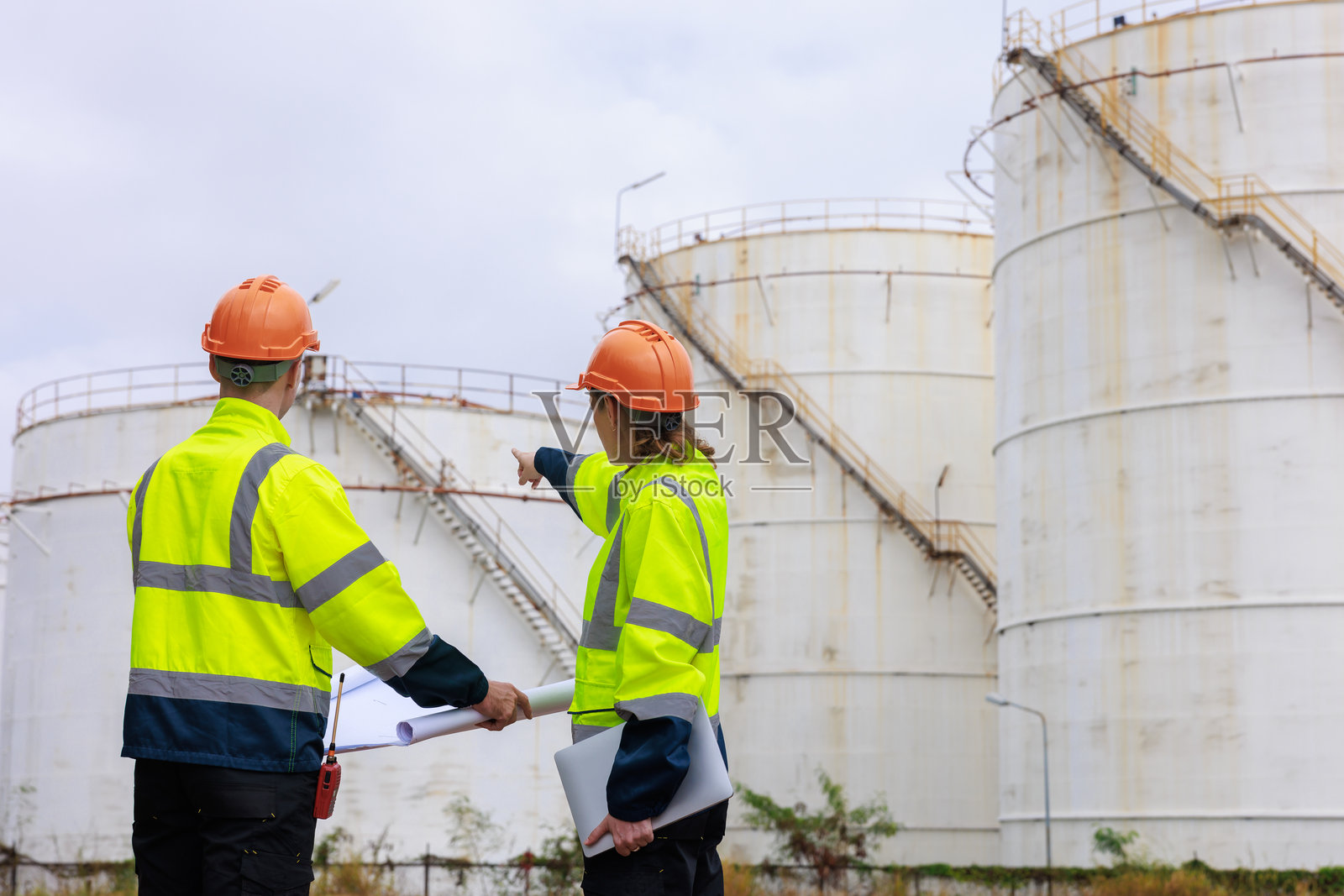 工程师测量团队穿着制服和安全帽，在工作场所检查蓝图项目，讨论和会面工作计划，背景是油储罐。照片摄影图片