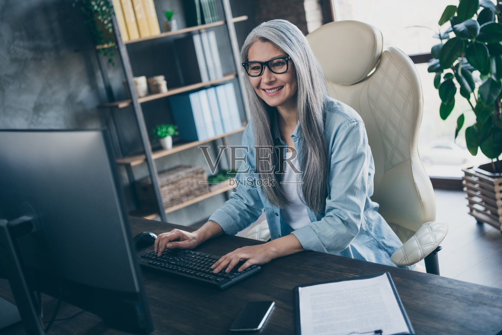 自信的退休女性金融家坐在桌前，通过电脑显示器进行视频通话，参加网络研讨会，工作空间内。照片摄影图片