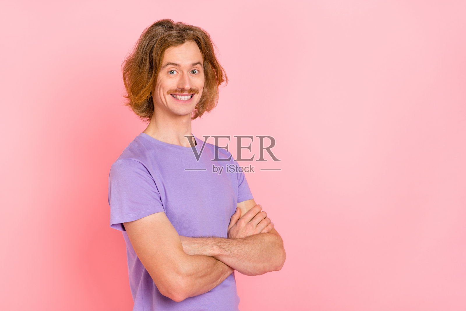 穿着紫色T恤的迷人快乐的时尚男士，双臂交叉，背景为粉色柔和色调，留有空白空间。照片摄影图片