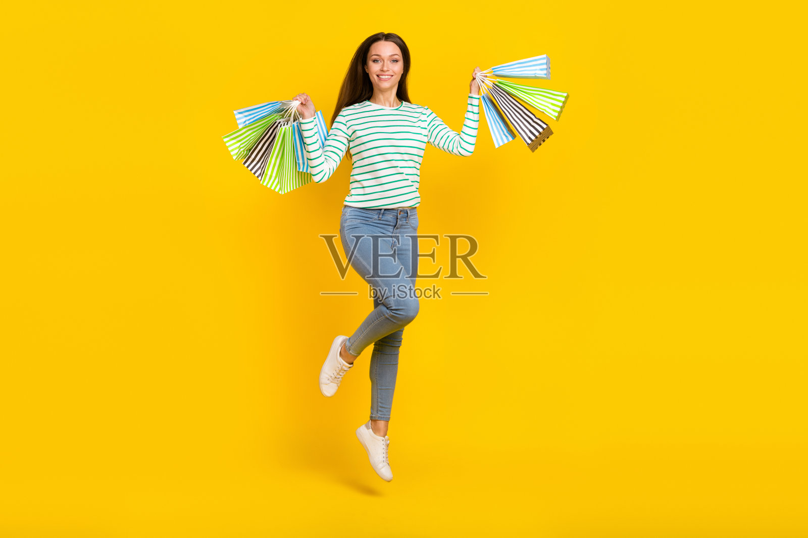 年轻女士跳起，手中提着购物袋，购买打折礼物，背景为黄色。照片摄影图片