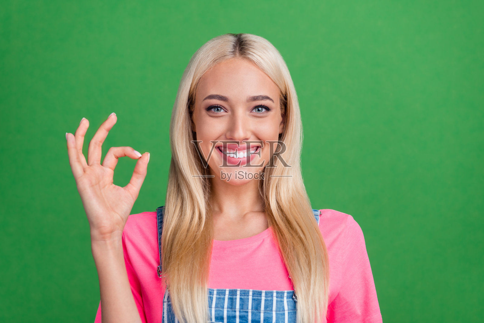 开心的女促销员展示“好”手势，赞同选择，穿着条纹工装，背景为绿色。照片摄影图片