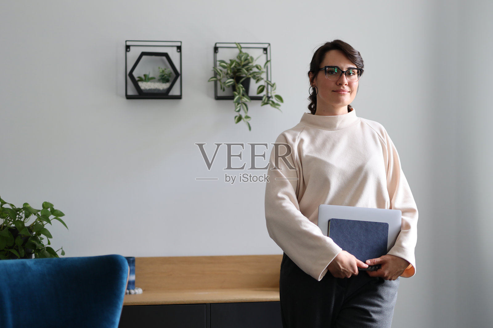 一位女性站在一个房间里，手边有一台笔记本电脑和一叠文件。她戴着眼镜，穿着白色衬衫。照片摄影图片