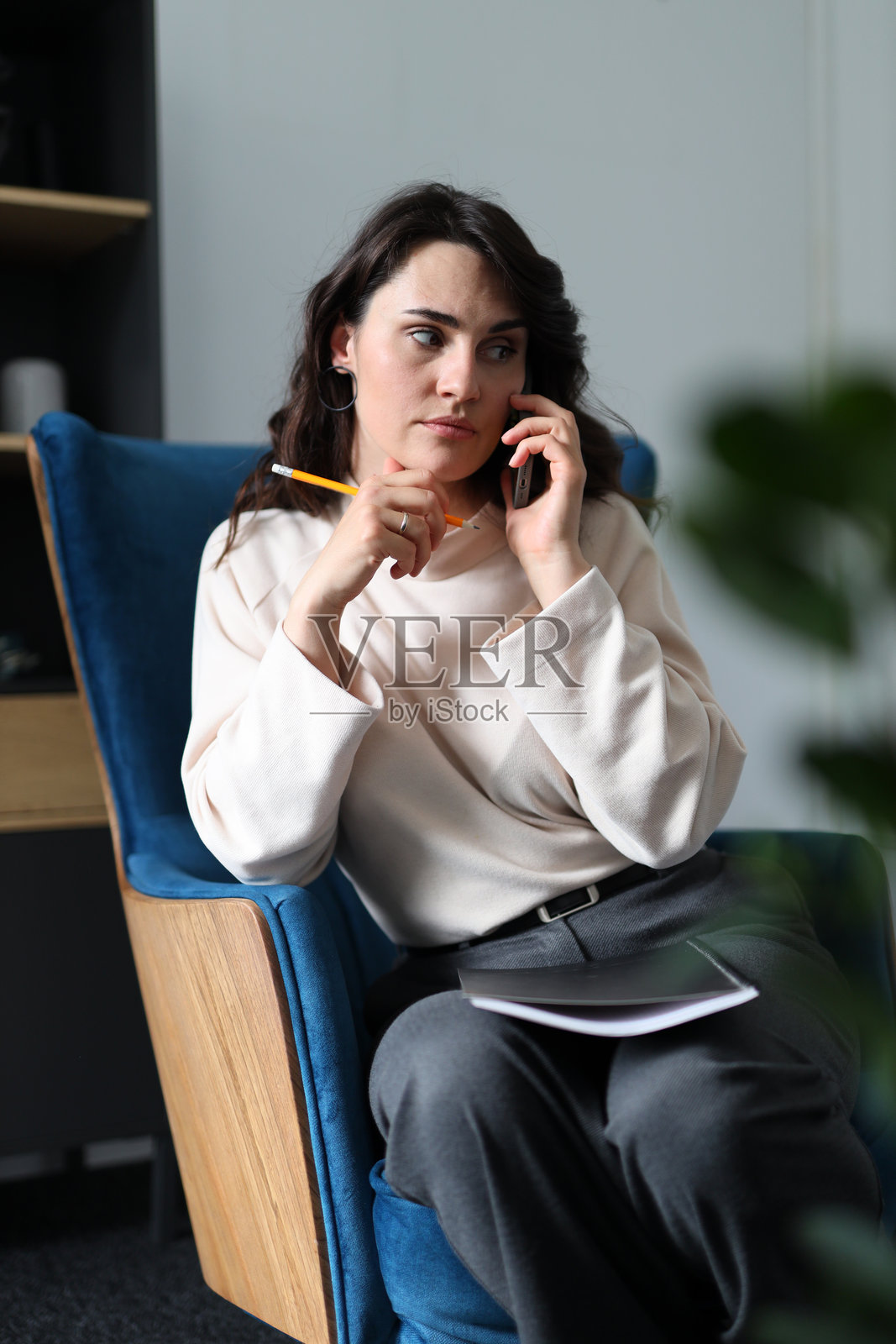 一位女性坐在蓝色椅子上，正在用手机通话。她穿着白色衬衫和灰色裤子。照片摄影图片