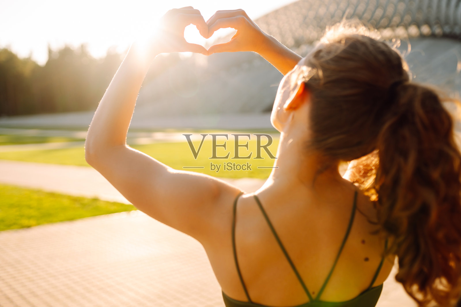 年轻女性穿着运动服在户外用双手做心形手势的背影。照片摄影图片