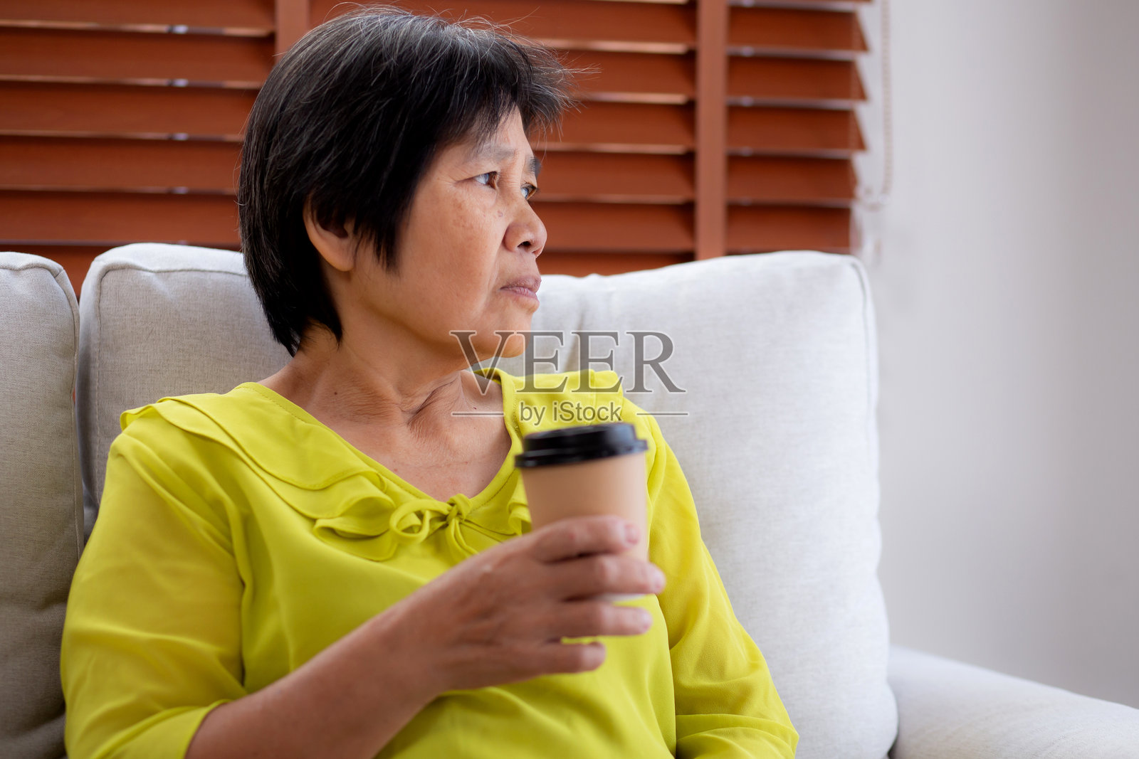 快乐的老年女性坐在沙发上，手握咖啡杯，望着窗外，享受家庭生活，悠闲地饮用饮料，体现生活方式和舒适感。照片摄影图片