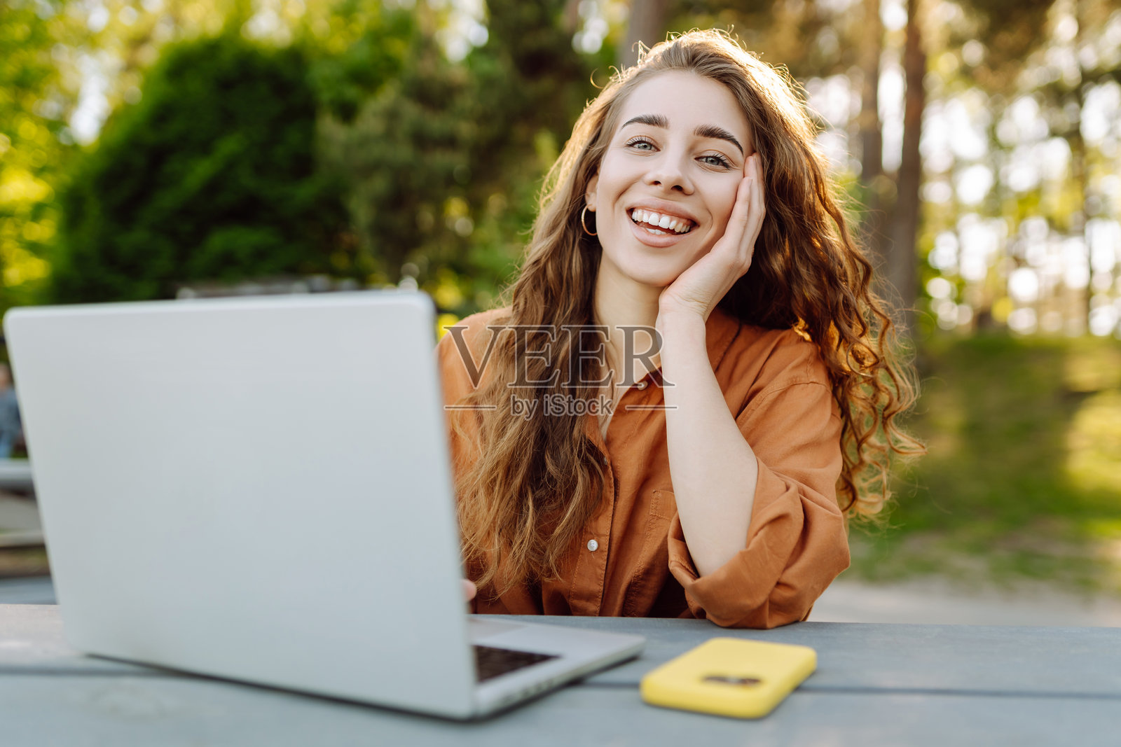 美丽的卷发女性在阳光明媚的公园里，坐在木桌前使用笔记本电脑工作。照片摄影图片