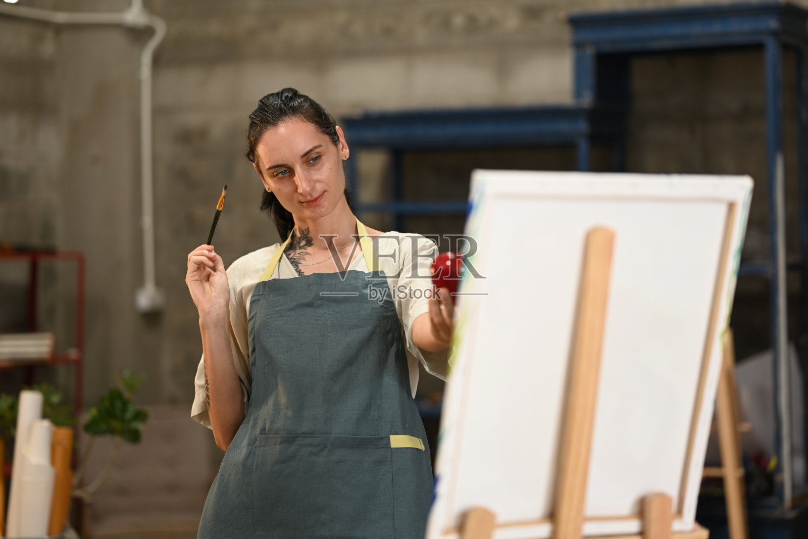 一位穿着围裙的白人女性艺术家在工作室中手握红苹果和画笔，准备将其本质描绘在画布上。照片摄影图片