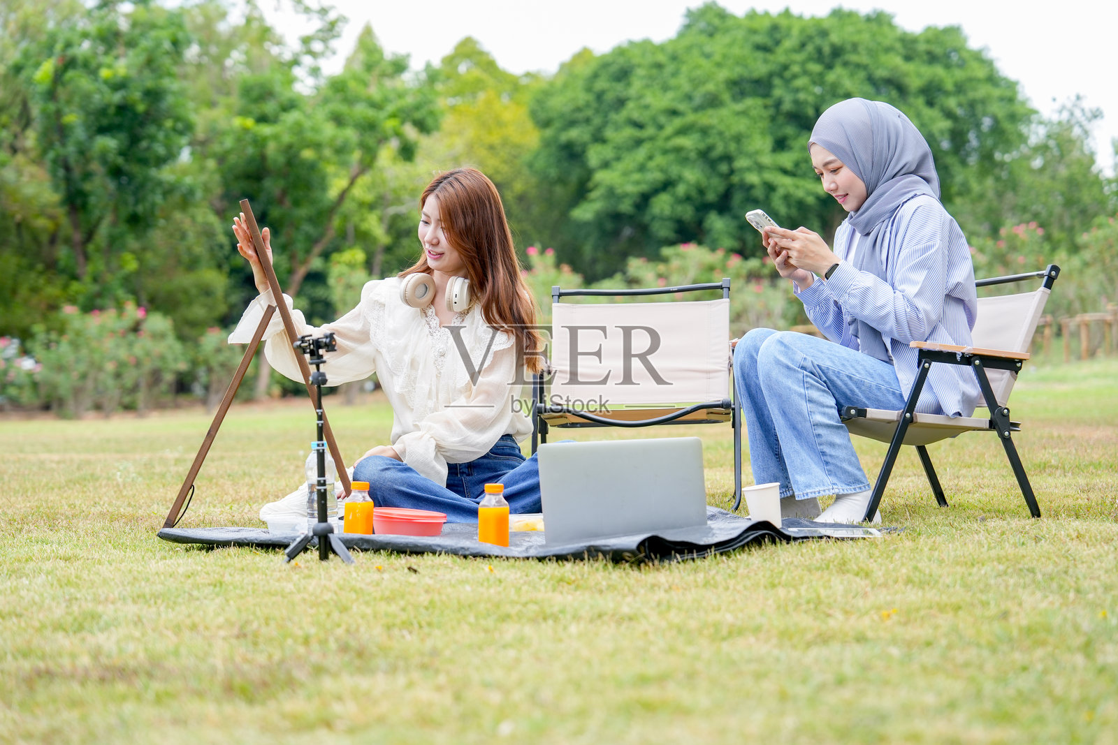 亚洲女性坐着准备黑板进行绘画，而穆斯林女性则坐在椅子上享受手机，在公共公园中。照片摄影图片