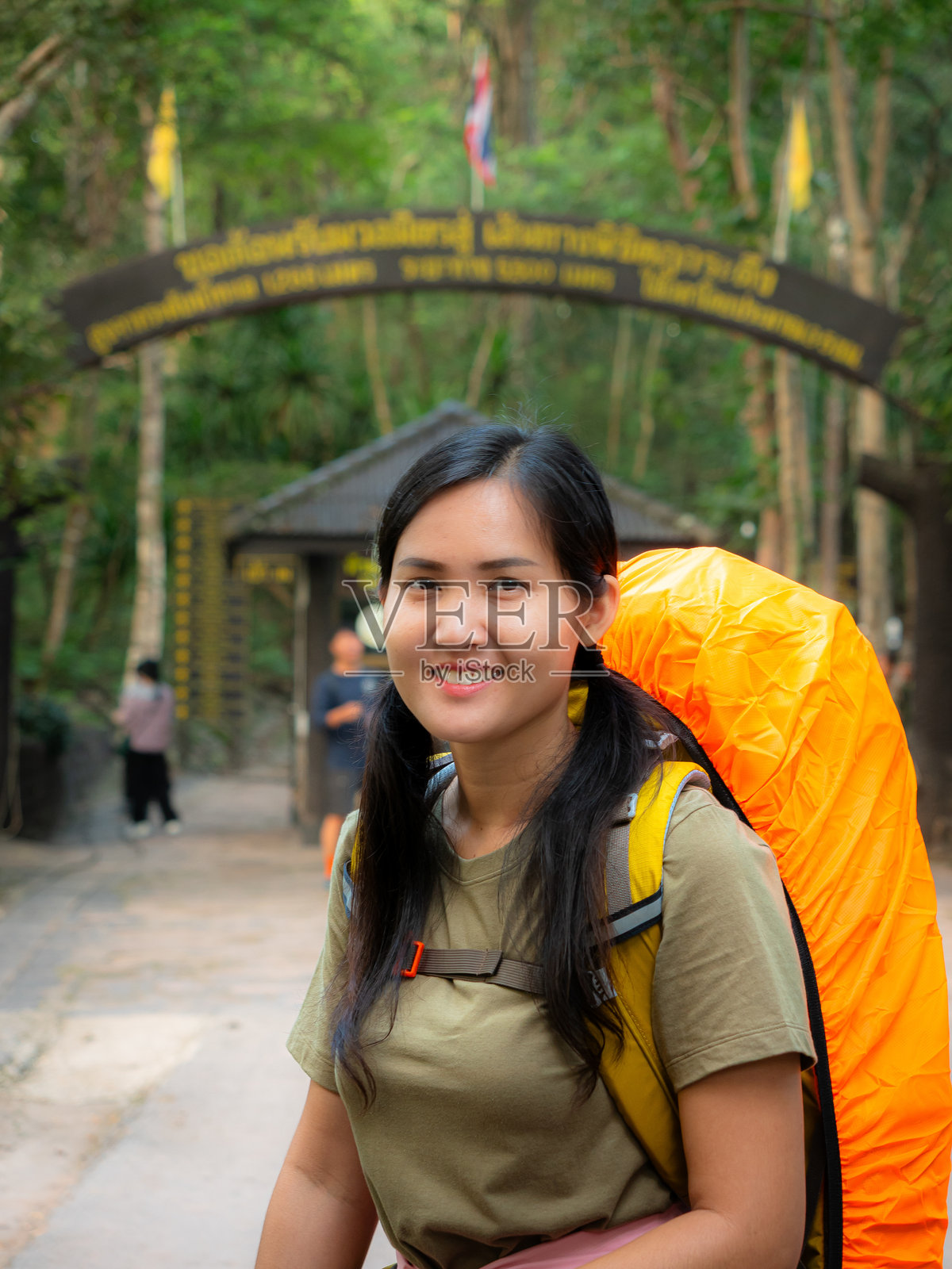 一位面带灿烂笑容的女性站在公园入口，背着一个大大的亮橙色背包，进行徒步旅行和户外活动。照片摄影图片