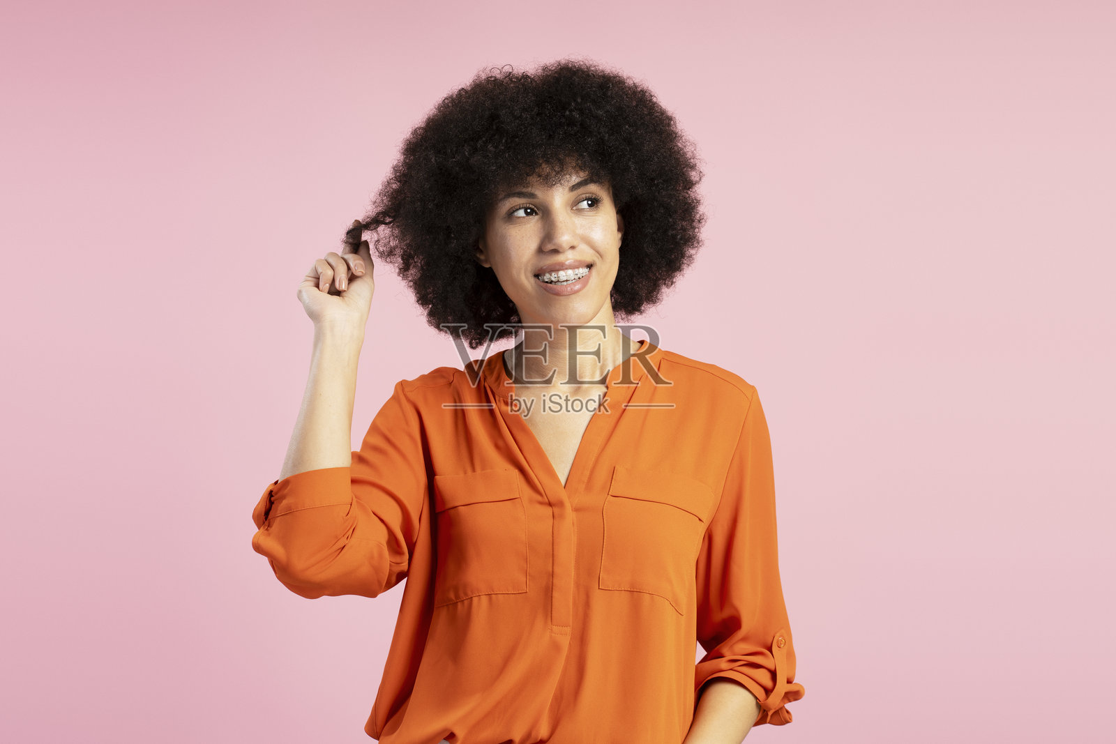 年轻的拉丁裔女性在粉色背景下玩弄头发，露出带牙套的微笑照片摄影图片