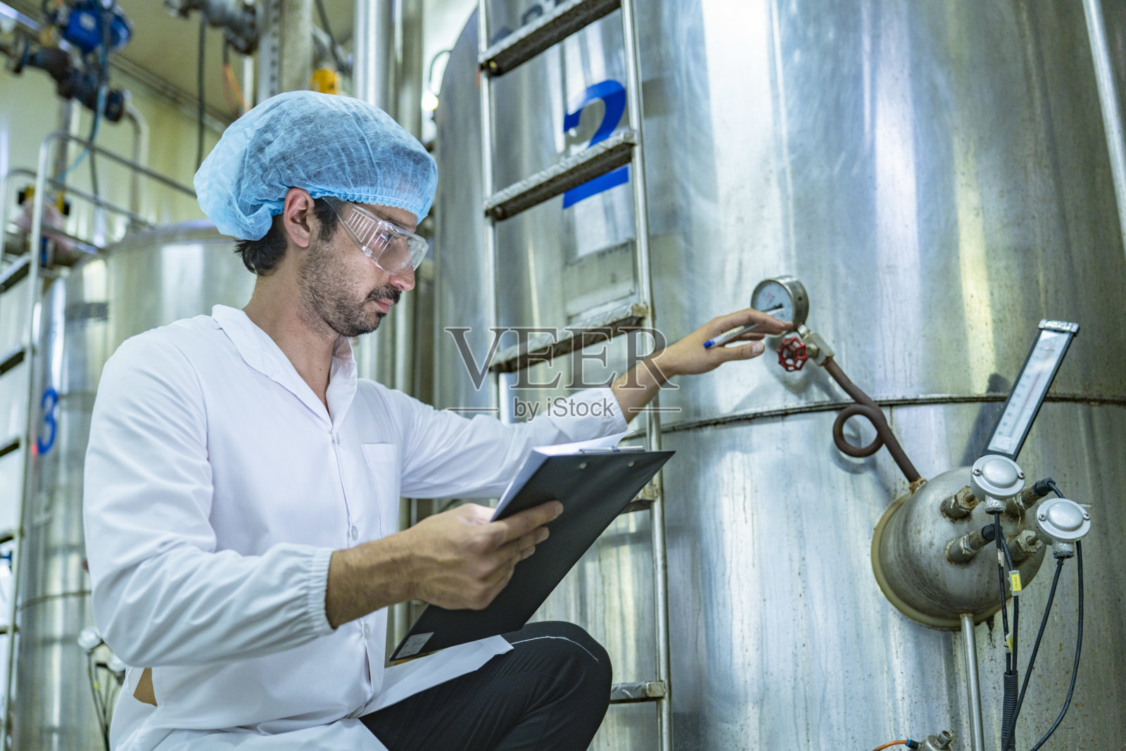 穿着白色工作服和安全装备的工人在检查大型工业罐，象征着质量控制、工业流程和制造操作中的精确性。照片摄影图片