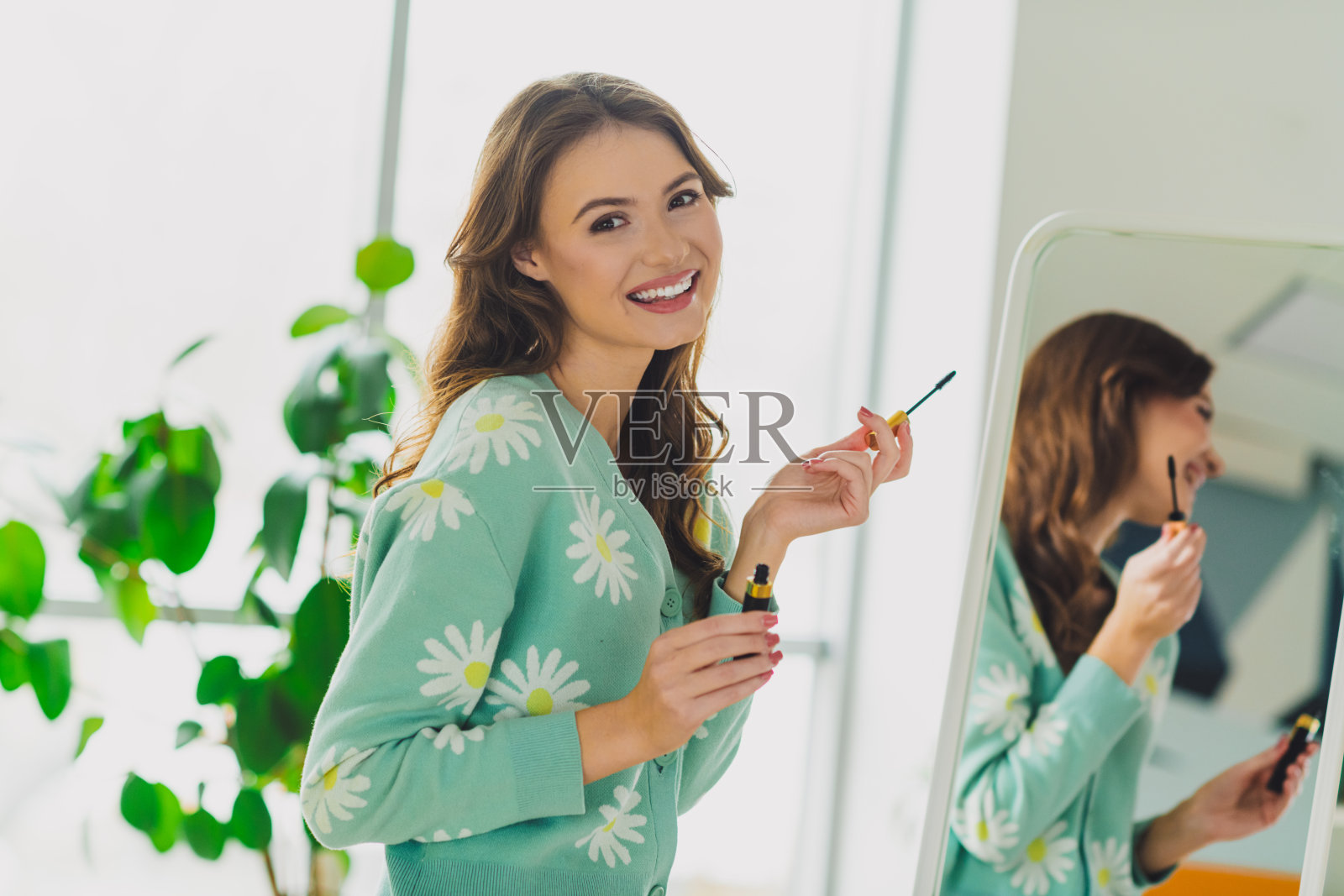 年轻女性在舒适的家居环境中微笑着涂抹睫毛膏，周围有时尚的装饰和自然的日光氛围。照片摄影图片