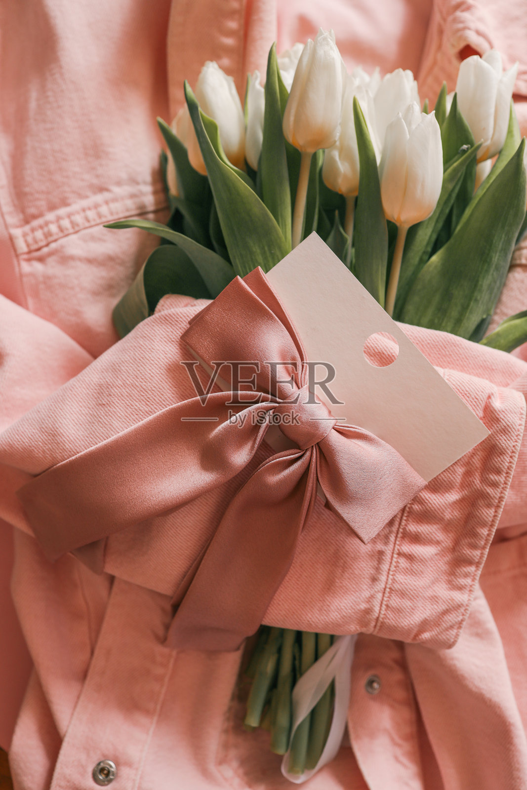 白色郁金香包裹在柔和的粉色牛仔夹克中，搭配缎面粉色发饰，摆放在卡片上，背景为柔和的粉色——一个迷人而精致的春季或庆祝活动平铺图。照片摄影图片
