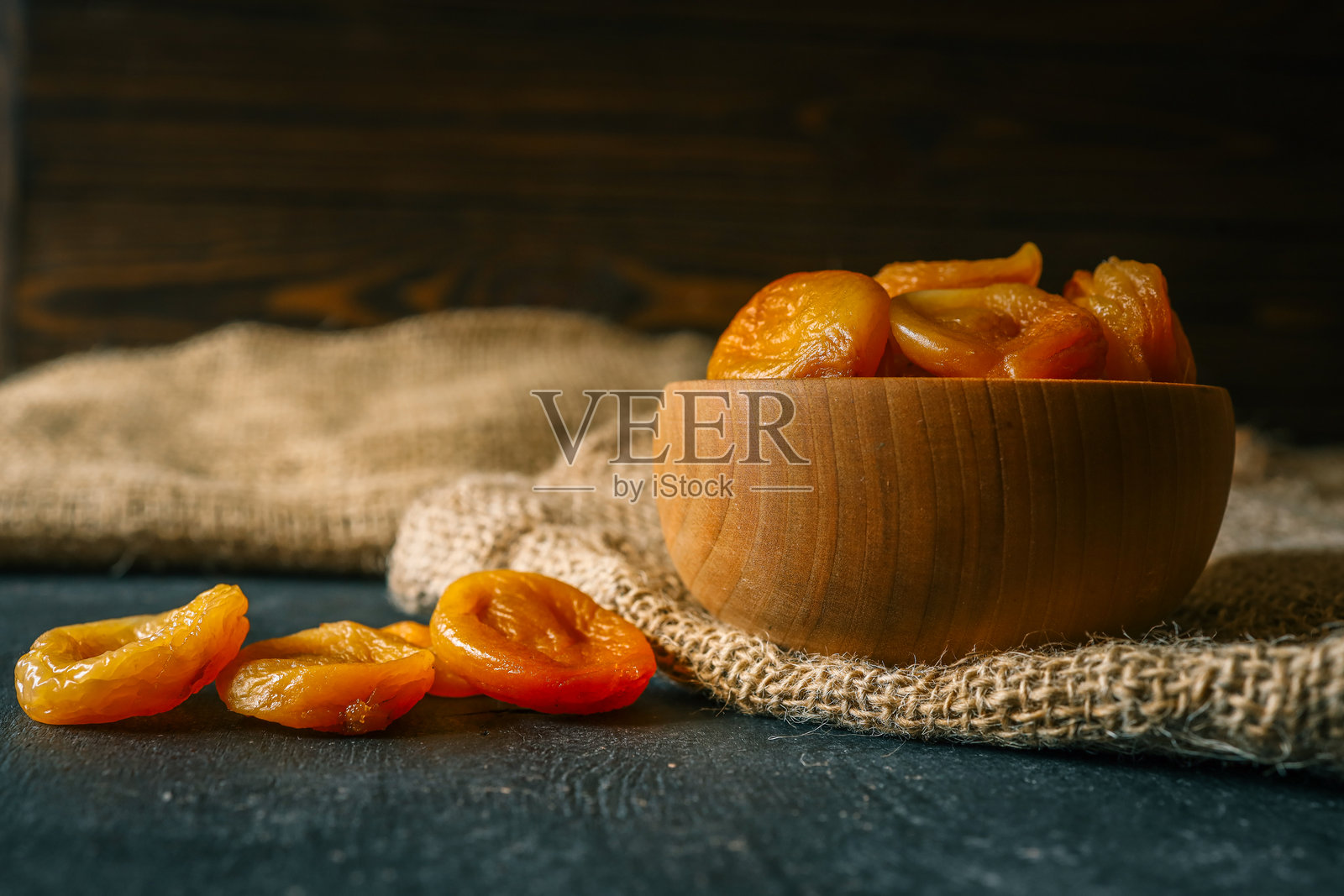 木碗和勺子里的干杏干，放在粗糙的麻布上。富含维生素和纤维的有机健康零食，在深色纹理背景下的自然温暖光线中拍摄。照片摄影图片