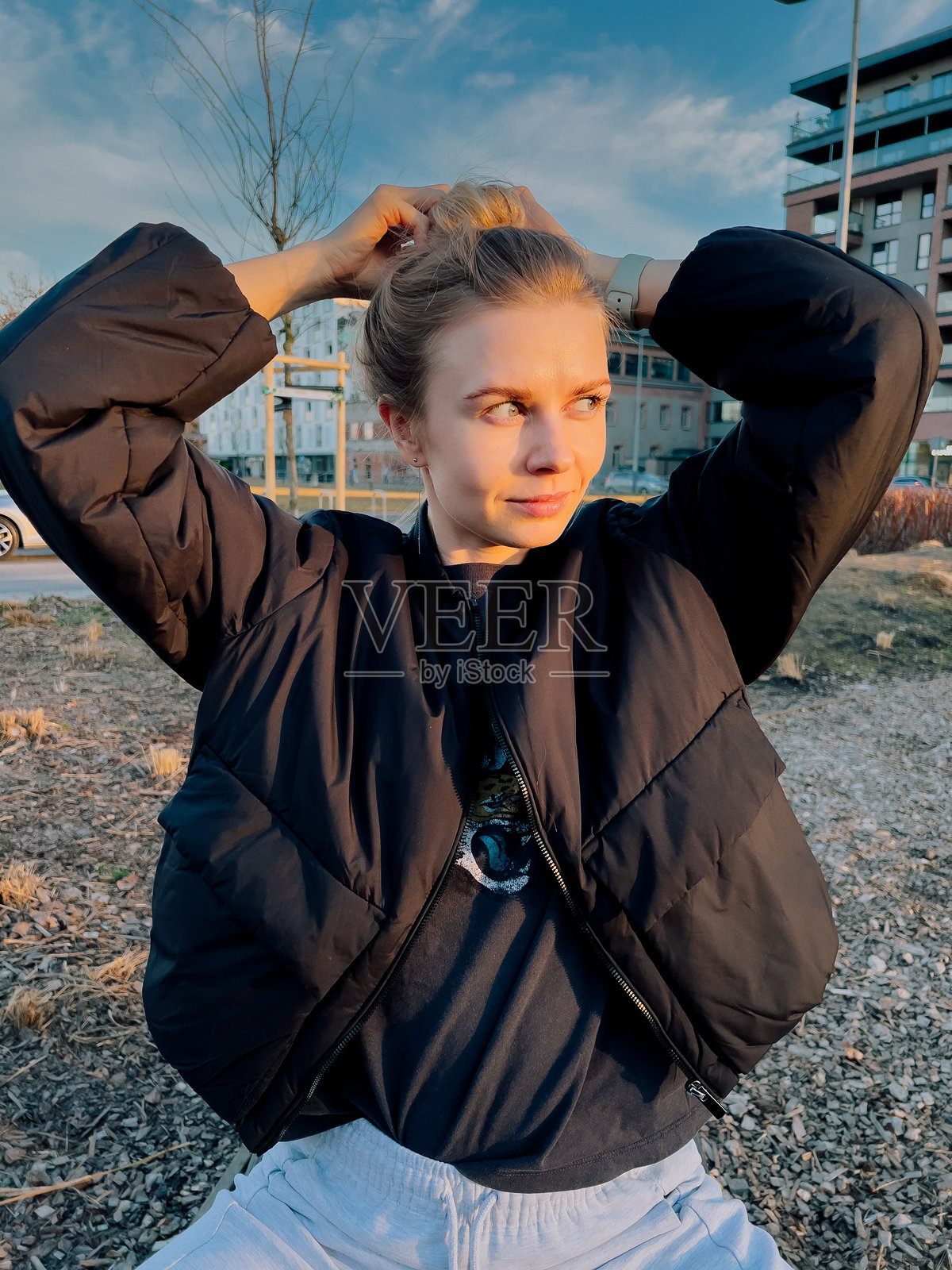 金发女子穿着黑色羽绒服和灰色运动裤，在城市公园的金色时光中享受阳光，正在把头发扎成发髻，营造出宁静而时尚的瞬间。照片摄影图片