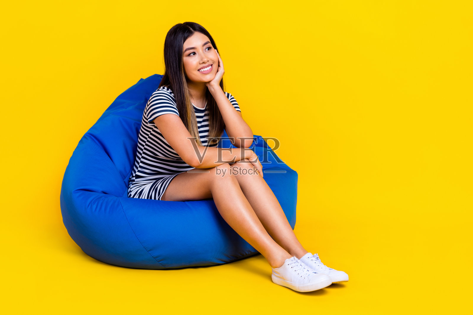 穿着条纹服装的梦幻甜美女性坐在豆袋椅上，凝视着空旷的空间，背景为黄色。照片摄影图片