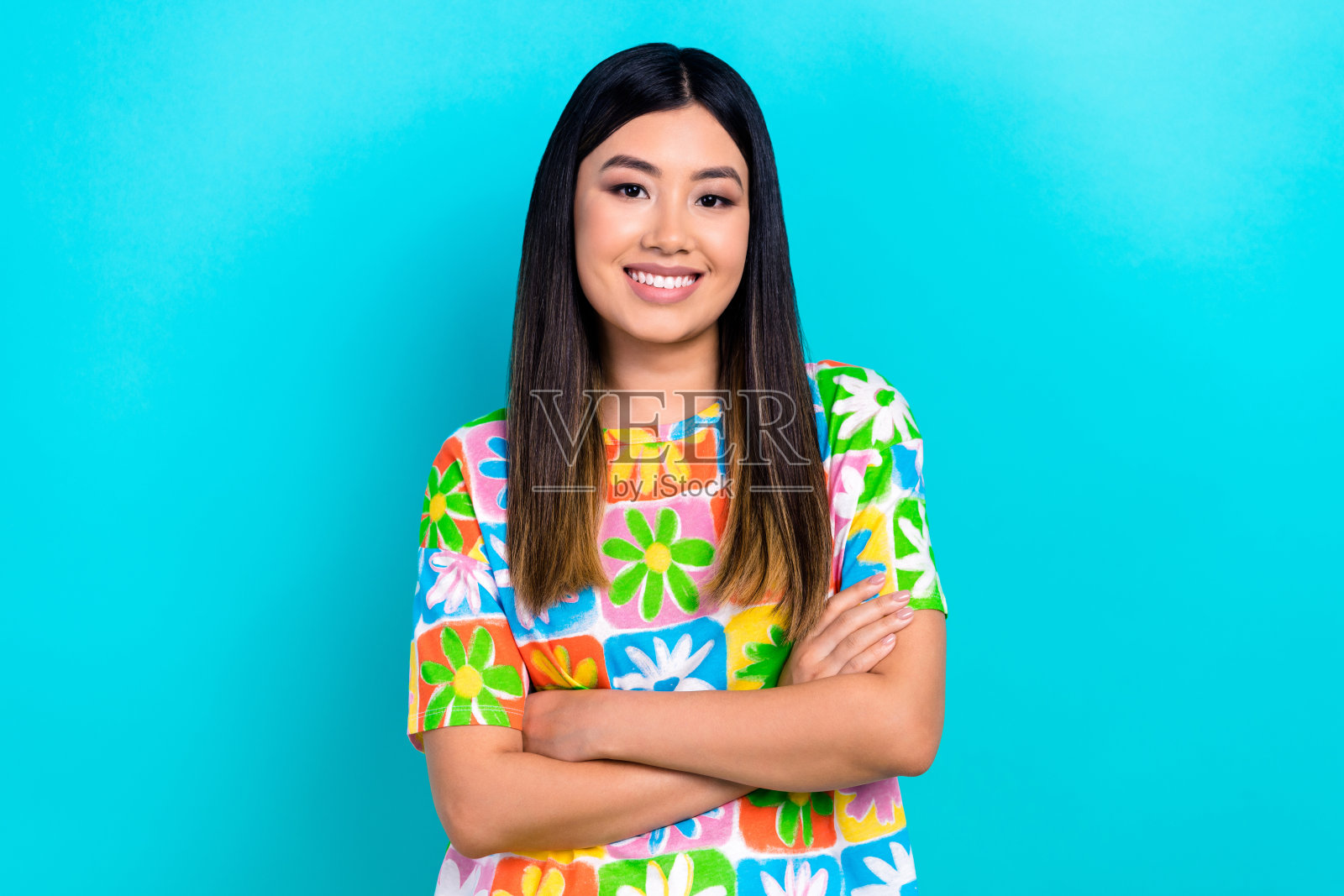 穿着花卉印花T恤的迷人积极女性微笑着，双臂交叉，背景为青色。照片摄影图片
