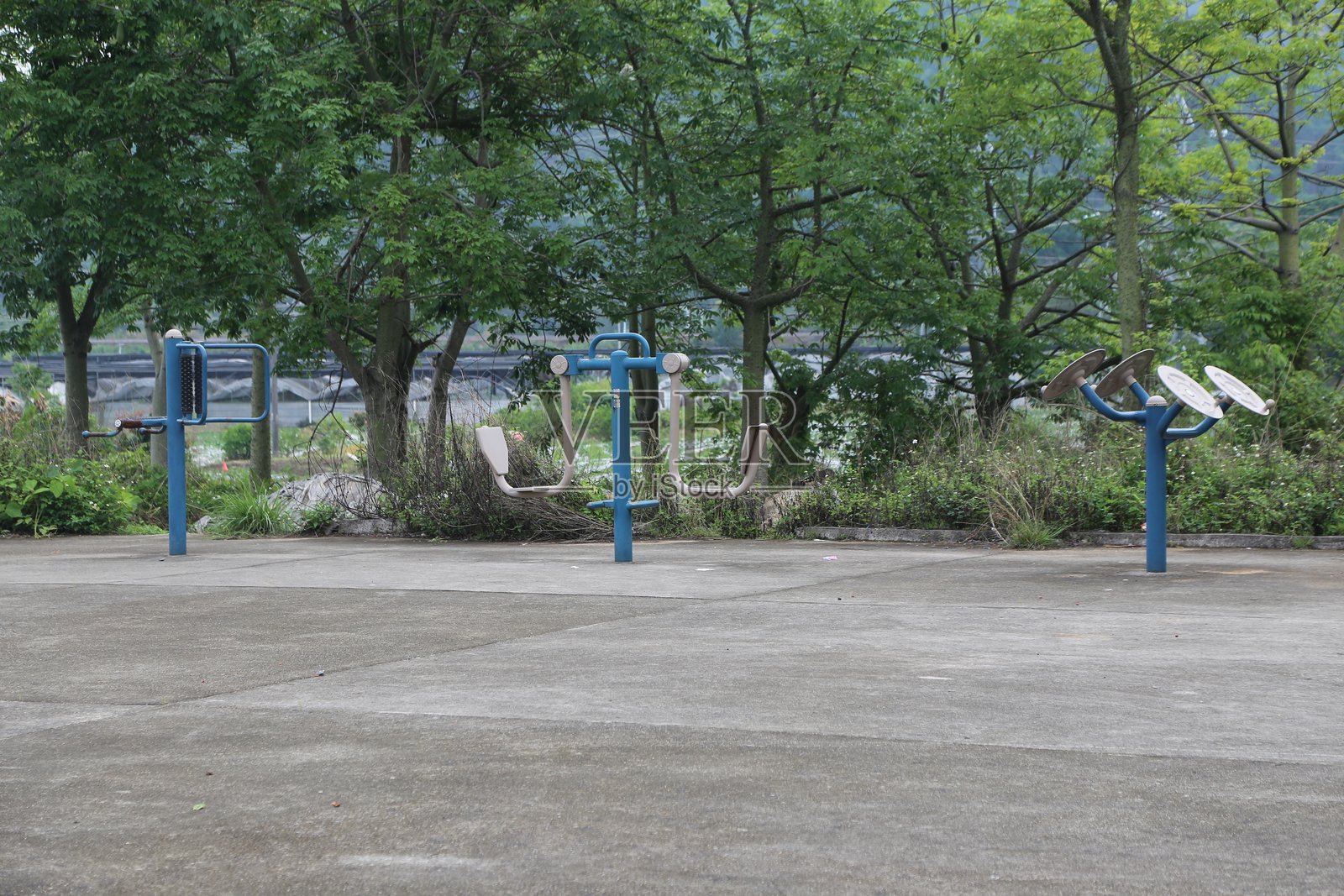 户外健身器材，蓝色金属健身设备，坐落在一个空旷的混凝土停车场，四周环绕着树木和绿植。照片摄影图片