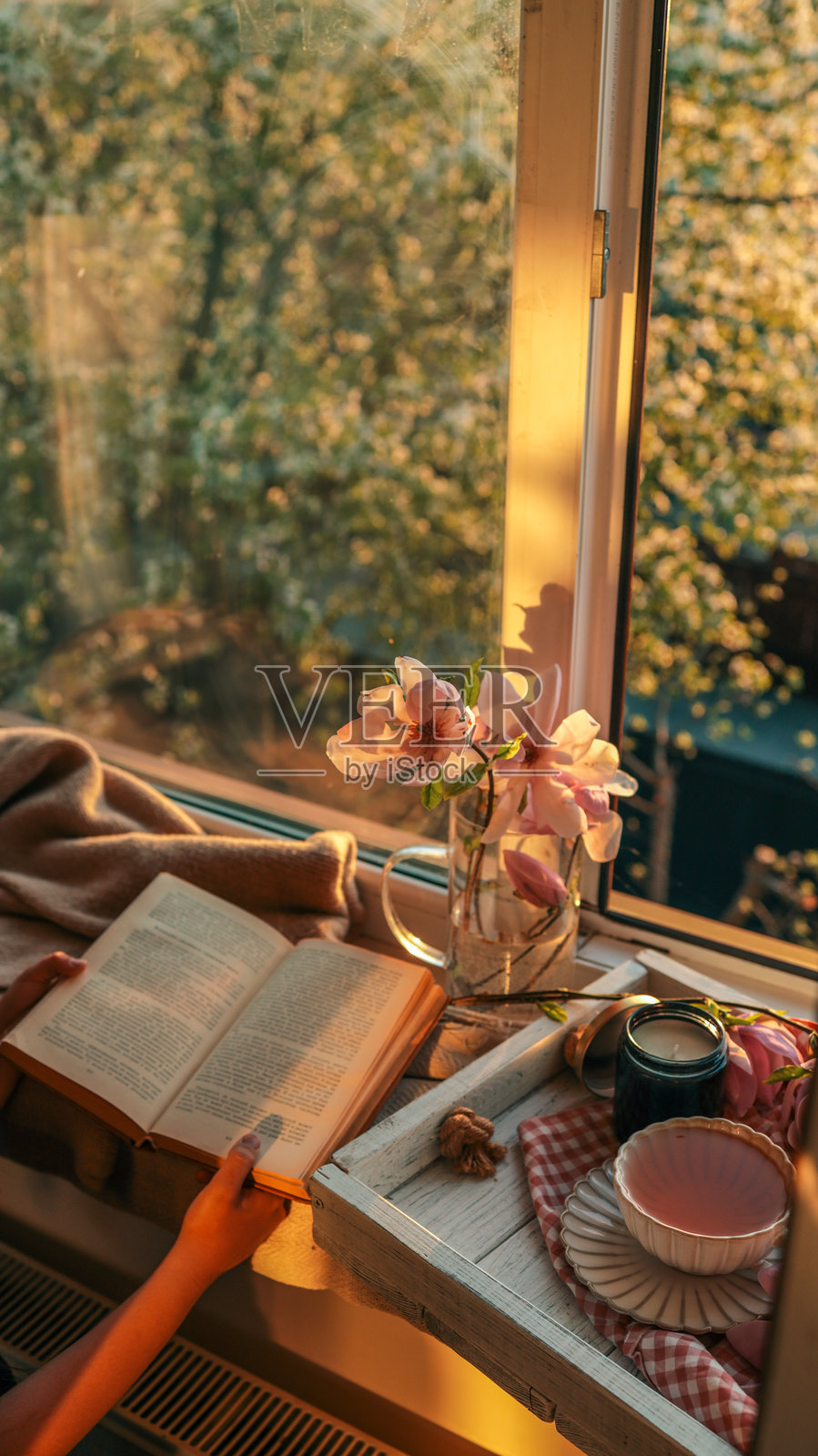 在温馨的窗边，夕阳下，一个人正阅读一本书，旁边有木兰花、一支黑色蜡烛和一杯茶放在木托盘上。温暖的金色光线营造出宁静、安详的氛围。照片摄影图片