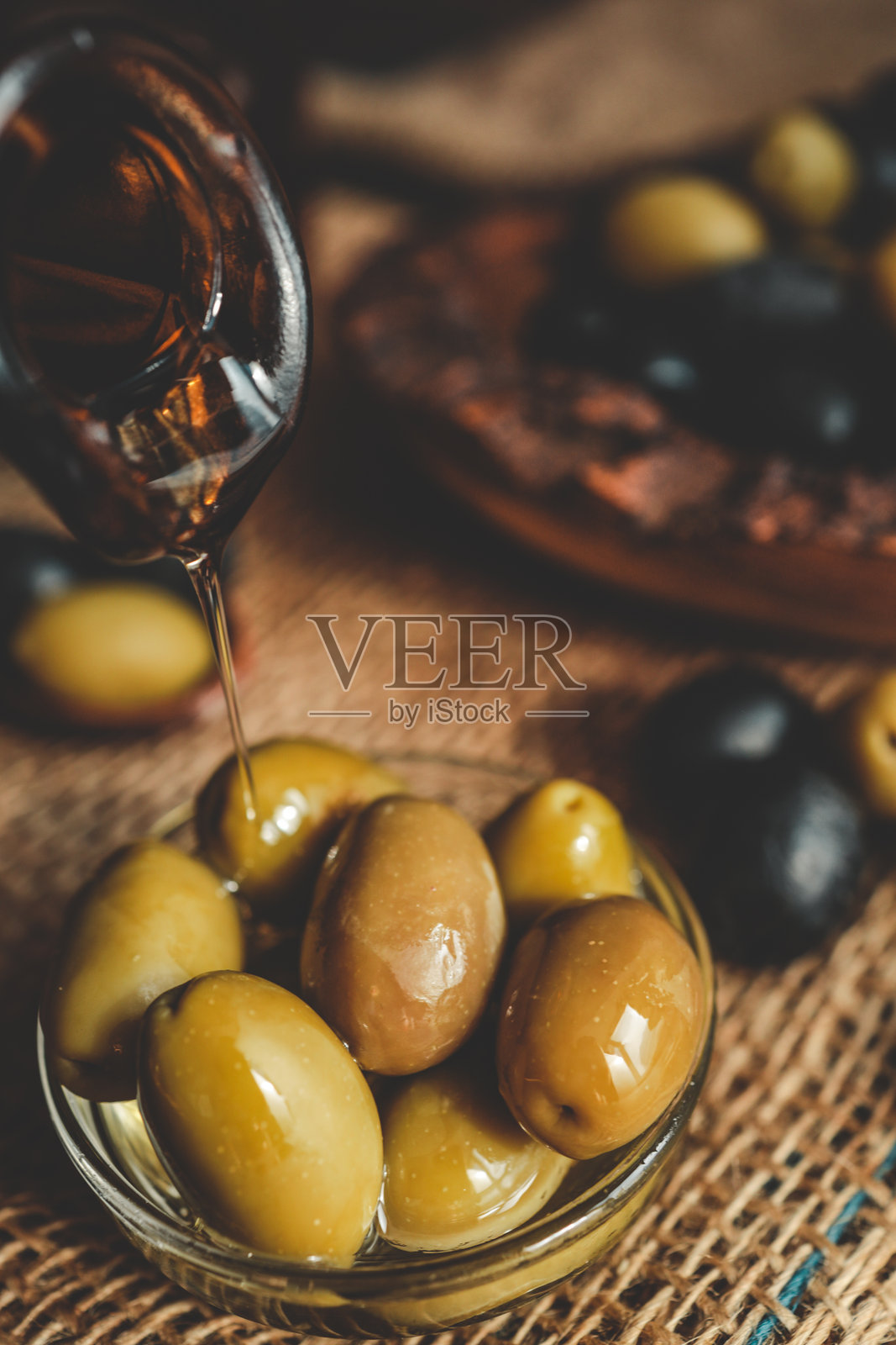 特写镜头：橄榄油倒在玻璃碗中的绿橄榄上，黑橄榄放在麻布上，背景是一个乡村风格的木碗，画面充满温暖的自然光。照片摄影图片