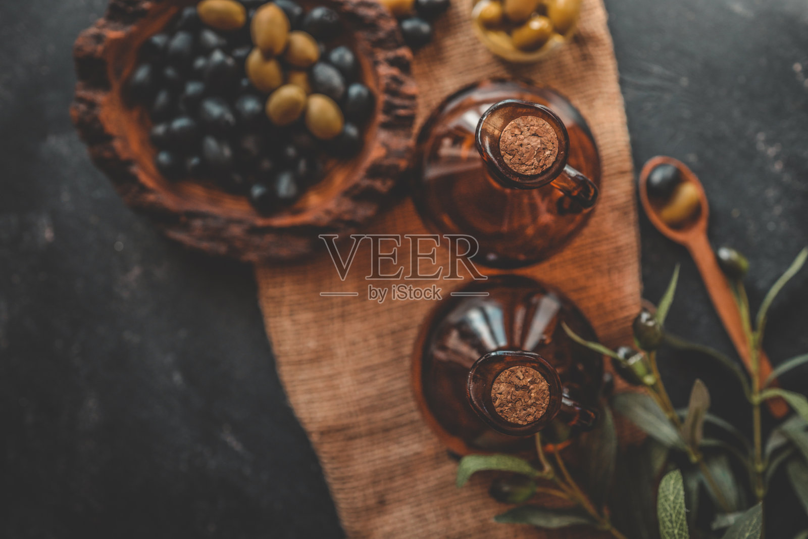 从上方俯视的乡村静物，包含绿色和黑色橄榄、玻璃瓶中的橄榄油、木碗和木勺，以及黑色纹理背景和麻布上的橄榄枝。照片摄影图片