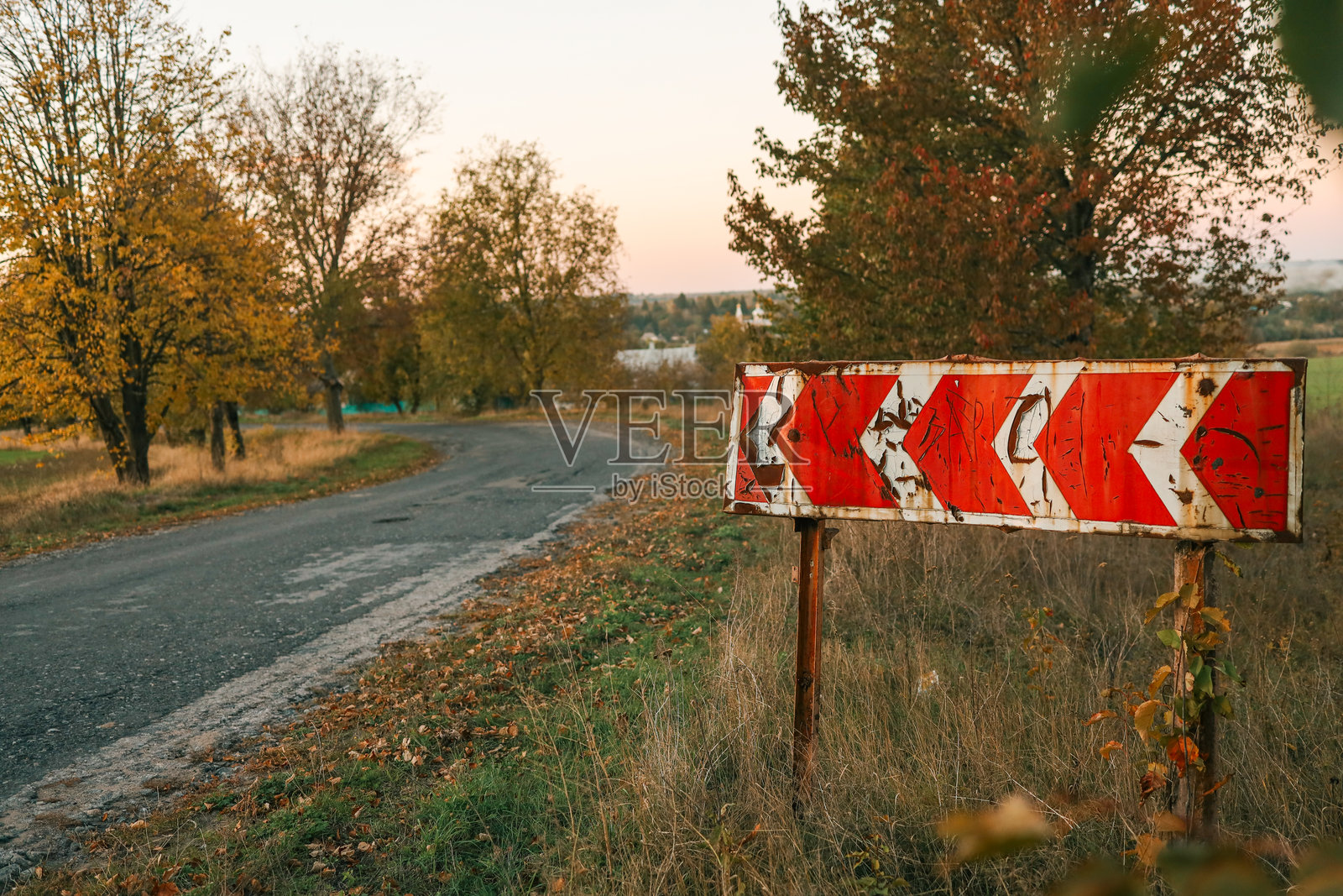 古老的风化路标，带有红色箭头，位于弯曲的乡村道路旁，周围是秋天的树木和干枯的草，在日落时分。照片摄影图片