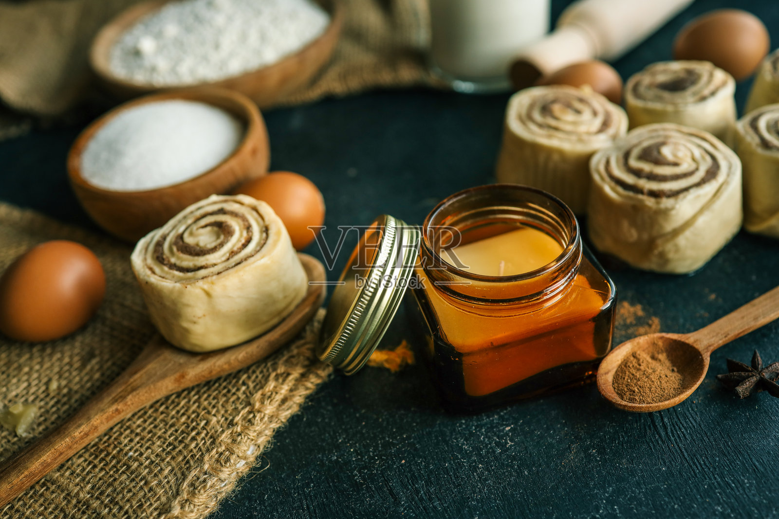 一个乡村风格的烘焙布置，包含肉桂卷、琥珀色蜡烛罐、面粉、糖和牛奶，放在深色表面上，完美适合温馨的厨房主题照片摄影图片