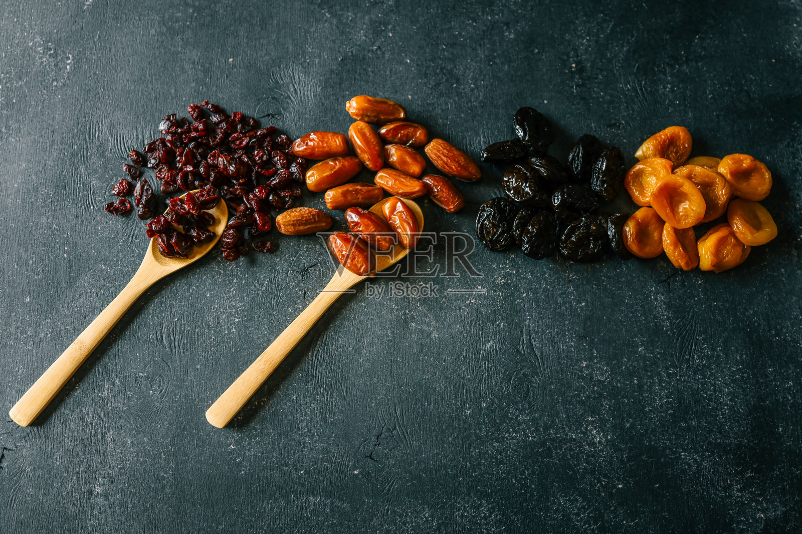 四种干果——蔓越莓、枣、梅干和杏干——在深色纹理背景上排列成行。色彩丰富且健康的天然零食组合。照片摄影图片