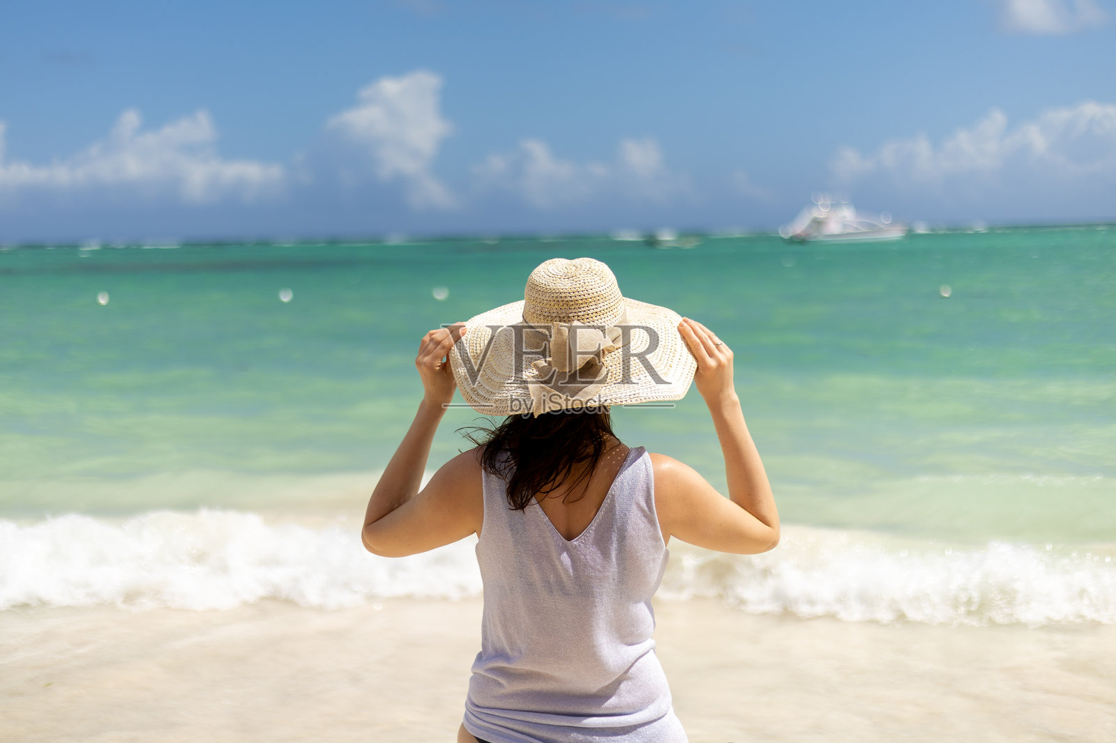 女人在清澈的蓝天下，戴着太阳帽站在浅水中，享受海滩美景。照片摄影图片