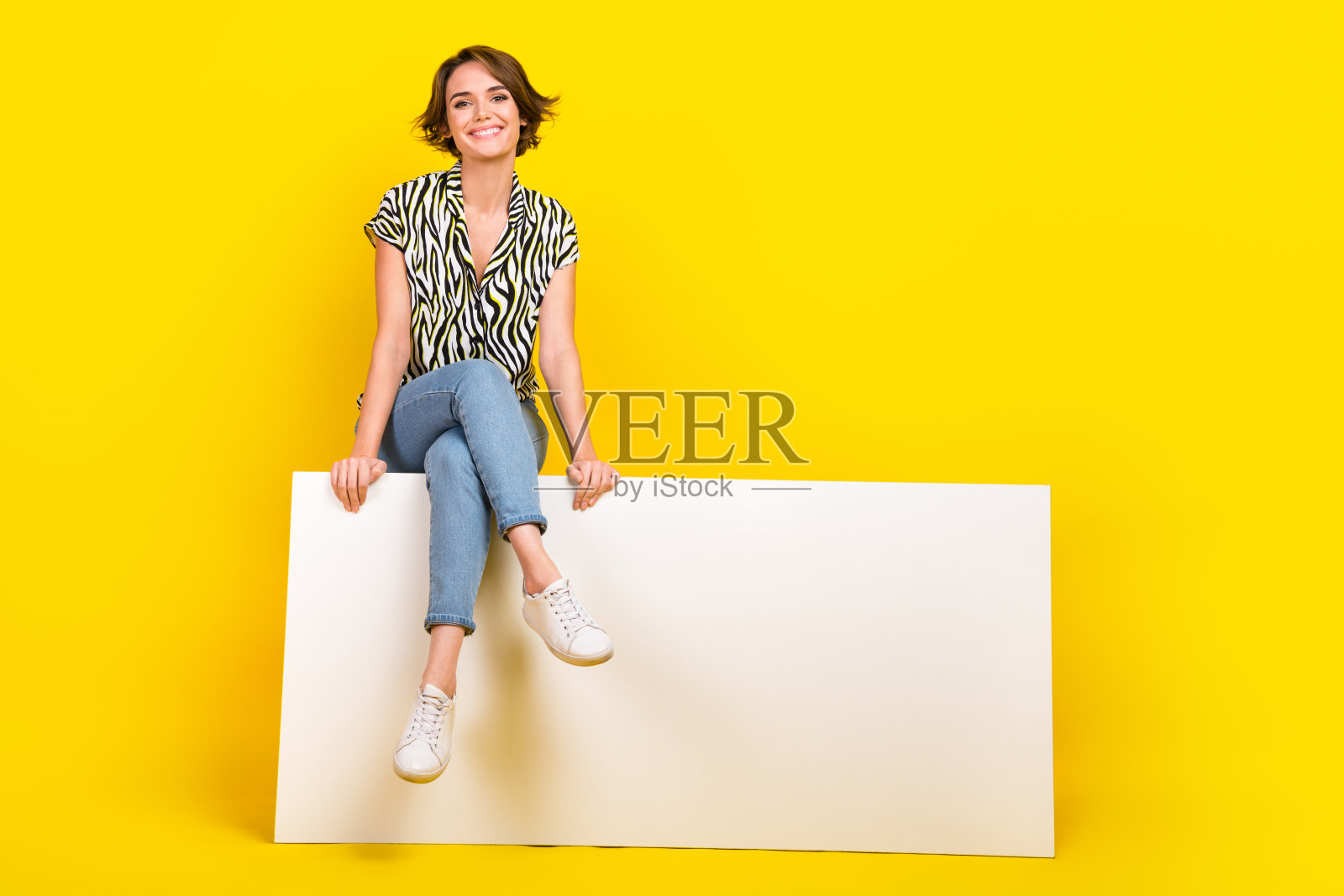 穿着斑马纹衬衫的可爱快乐女性全身照，坐在空白标牌前，背景为黄色。照片摄影图片