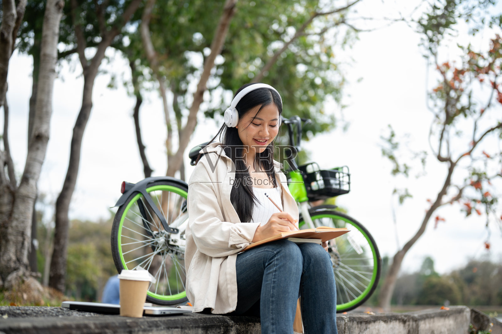 一位亚洲女性戴着耳机，愉快地在笔记本上写字，坐在公园的人行道上。照片摄影图片