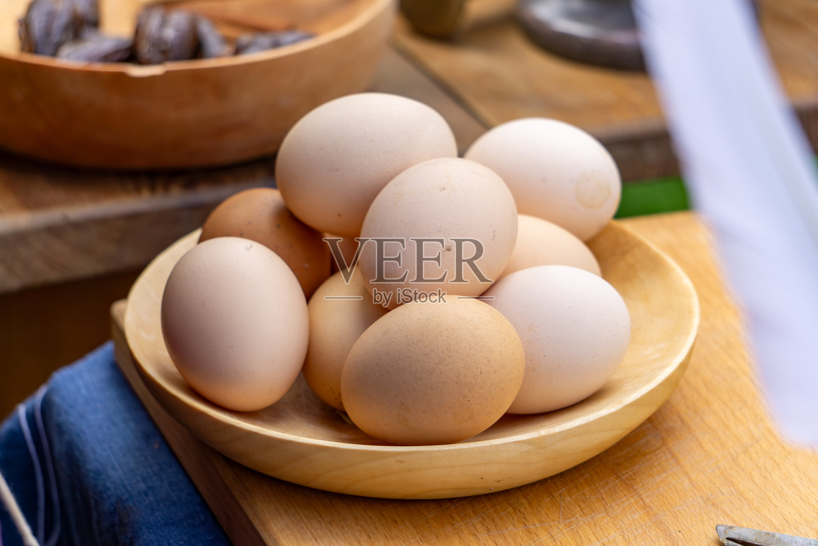 一个装满新鲜鸡蛋的木碗放在桌子上，背景中还有其他食物。这是一个自然、乡村风格的摆放，完美地宣传健康食品和农产品。照片摄影图片