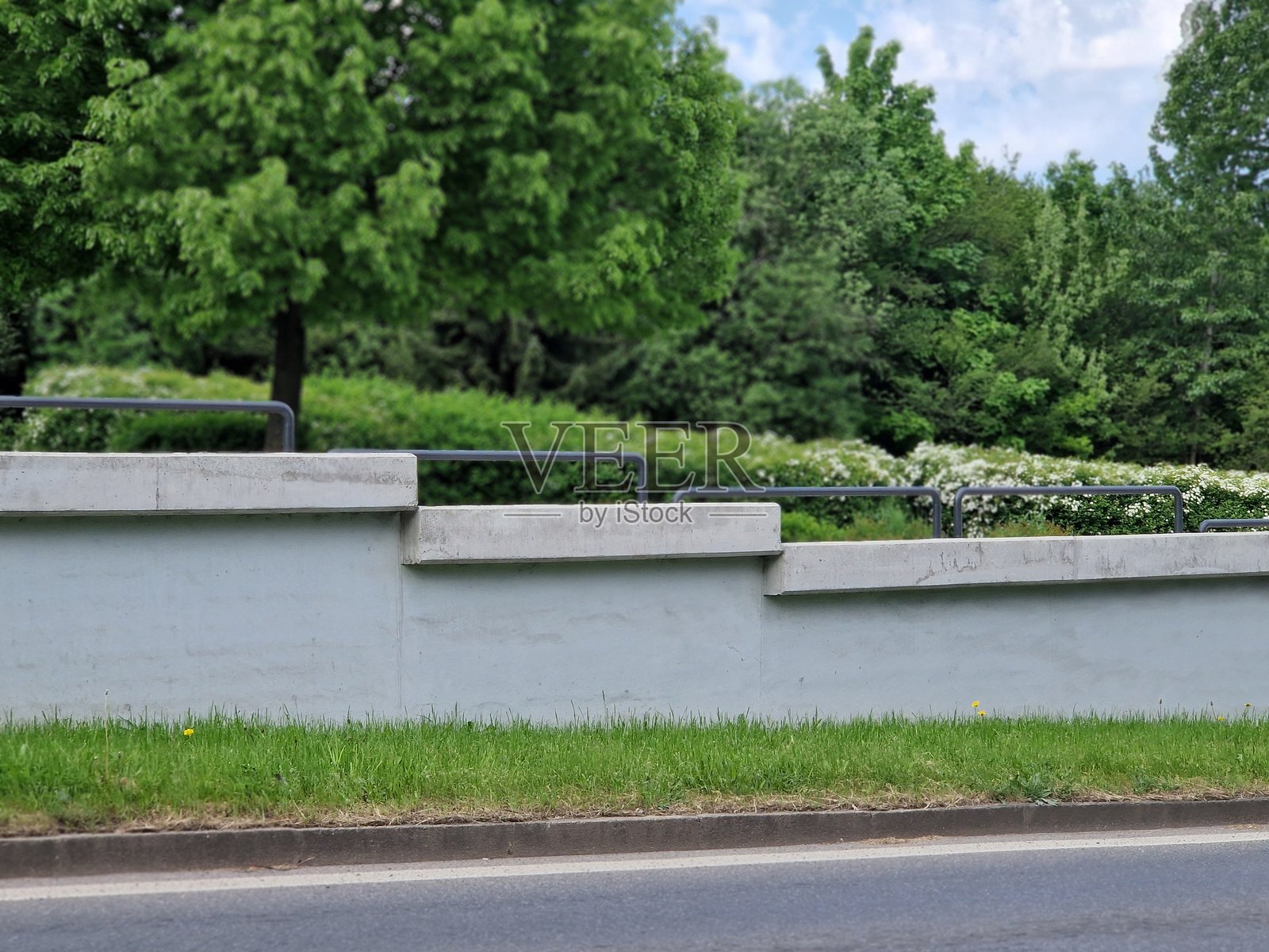 路边的大型混凝土挡土墙，配有管状栏杆。背景中有开花的灌木、树木和公园。照片摄影图片